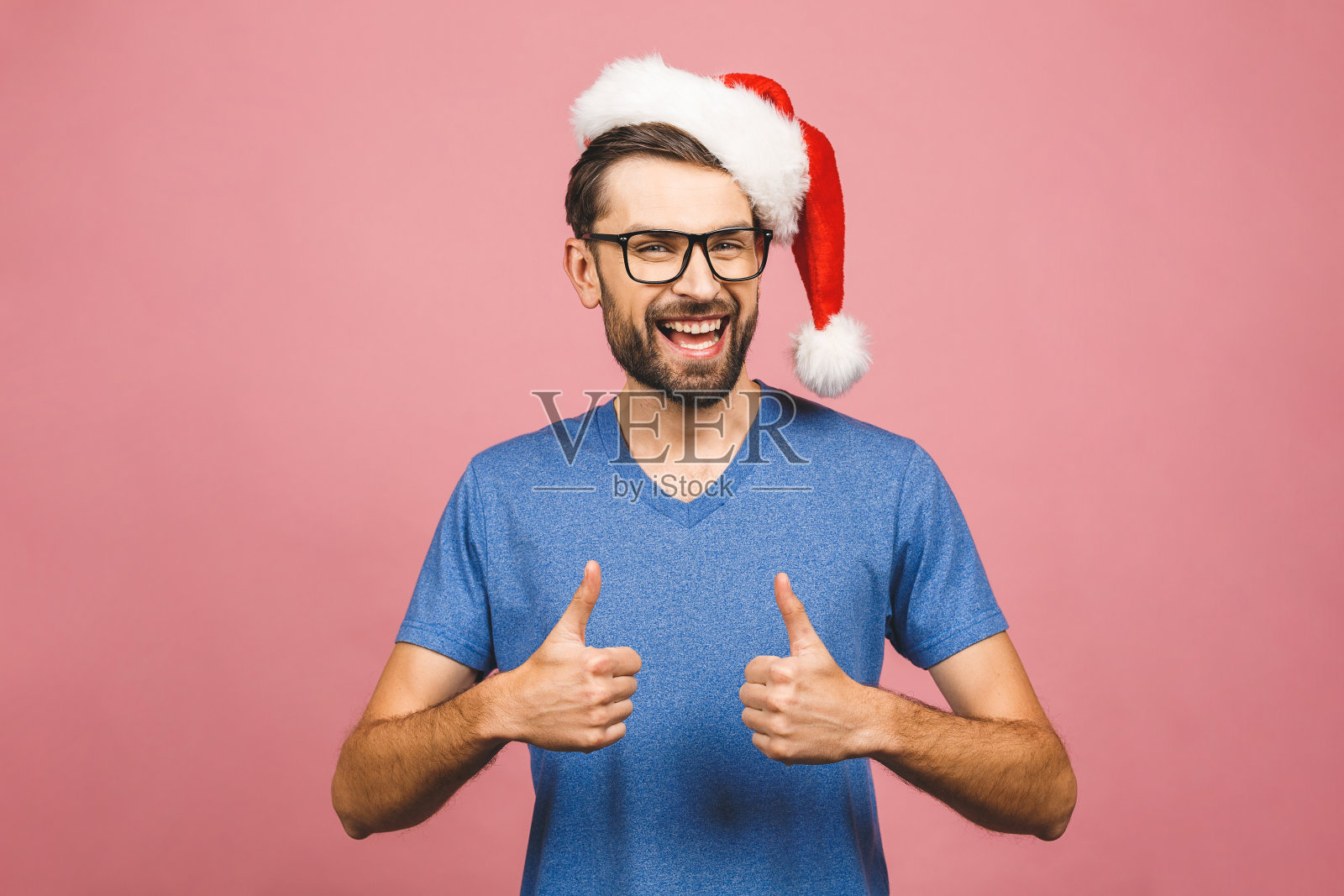 年轻英俊的男子戴着圣诞帽孤立粉红色的背景微笑积极。成功的表达。竖起大拇指。照片摄影图片