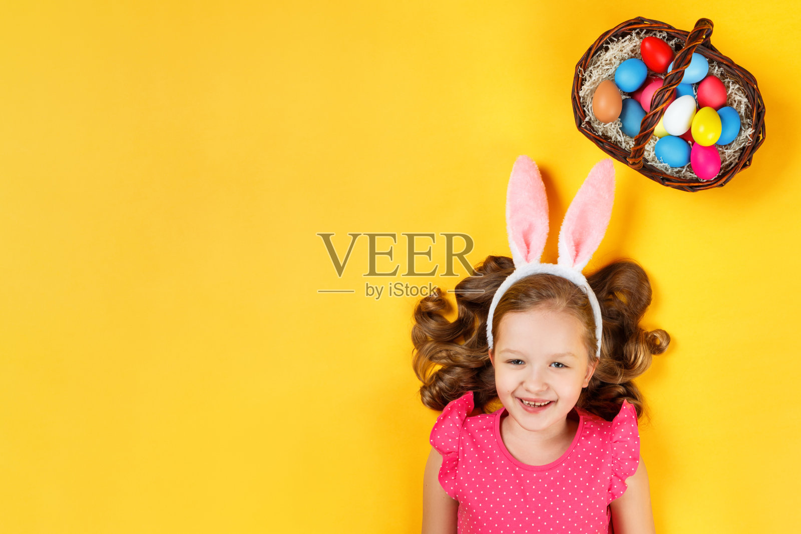 俯视图上一个快乐的婴儿在兔子的耳朵和一个篮子与复活节彩蛋。一个小女孩的肖像躺在地板上的黄色背景照片摄影图片