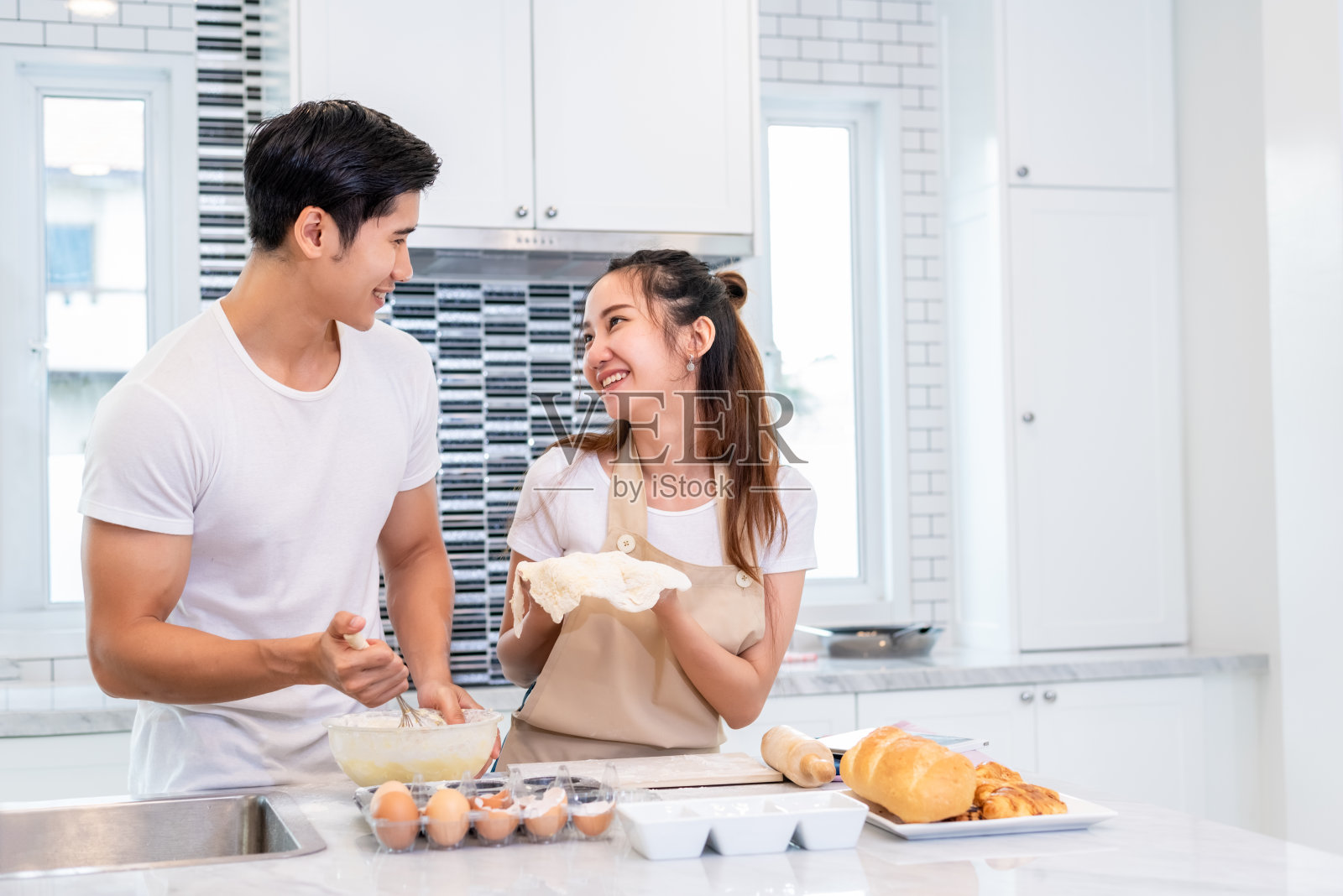快乐的亚洲夫妇一起在厨房做饭和烤蛋糕。男女望着药片在家遵循食谱步骤。爱和幸福的概念。甜蜜的蜜月和情人节主题照片摄影图片
