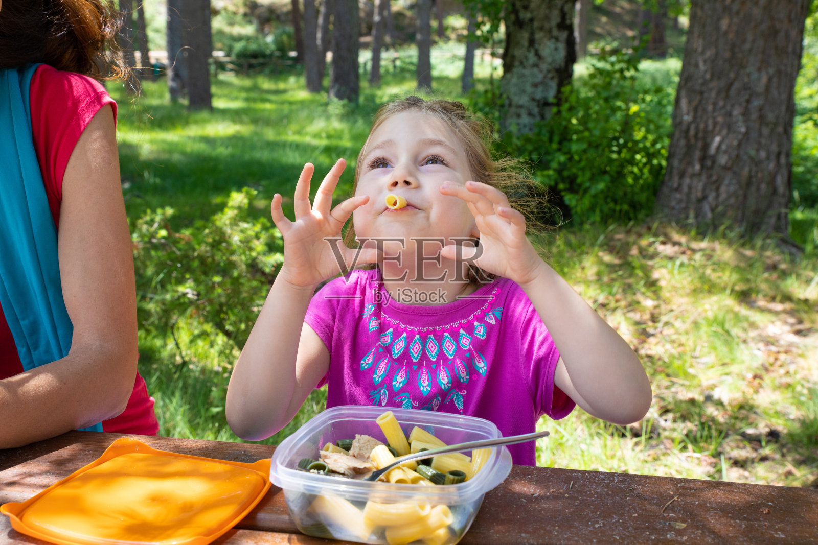 有趣的小女孩玩通心粉吃塑料午餐盒旁边她的母亲在餐桌野餐照片摄影图片