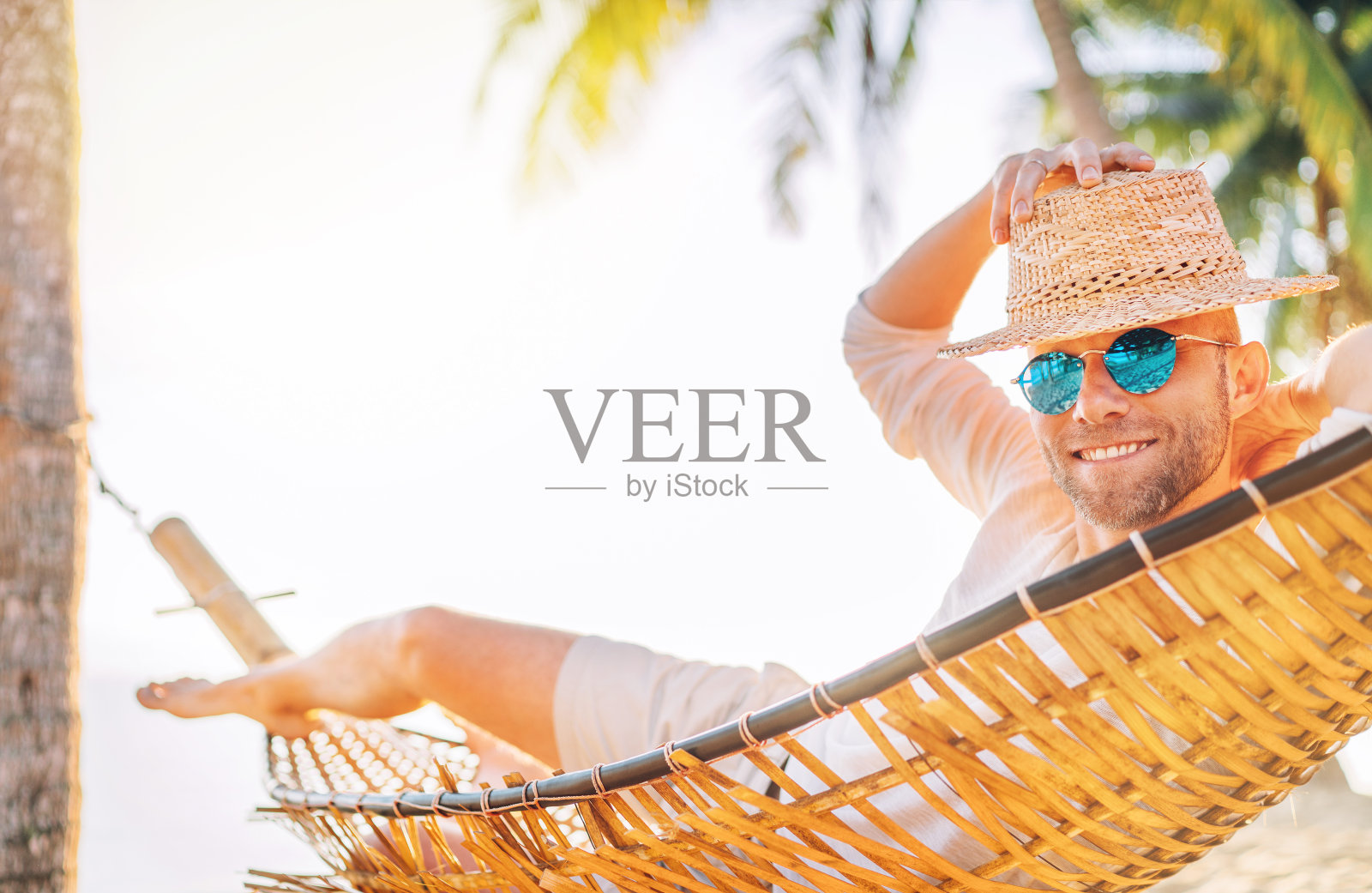 戴着草帽和蓝色太阳镜的年轻人躺在舒适的吊床上晃荡，真诚地微笑着迎接早晨的日出阳光在沙滩上的棕榈树下。平静异国风情的地方度假概念形象。照片摄影图片