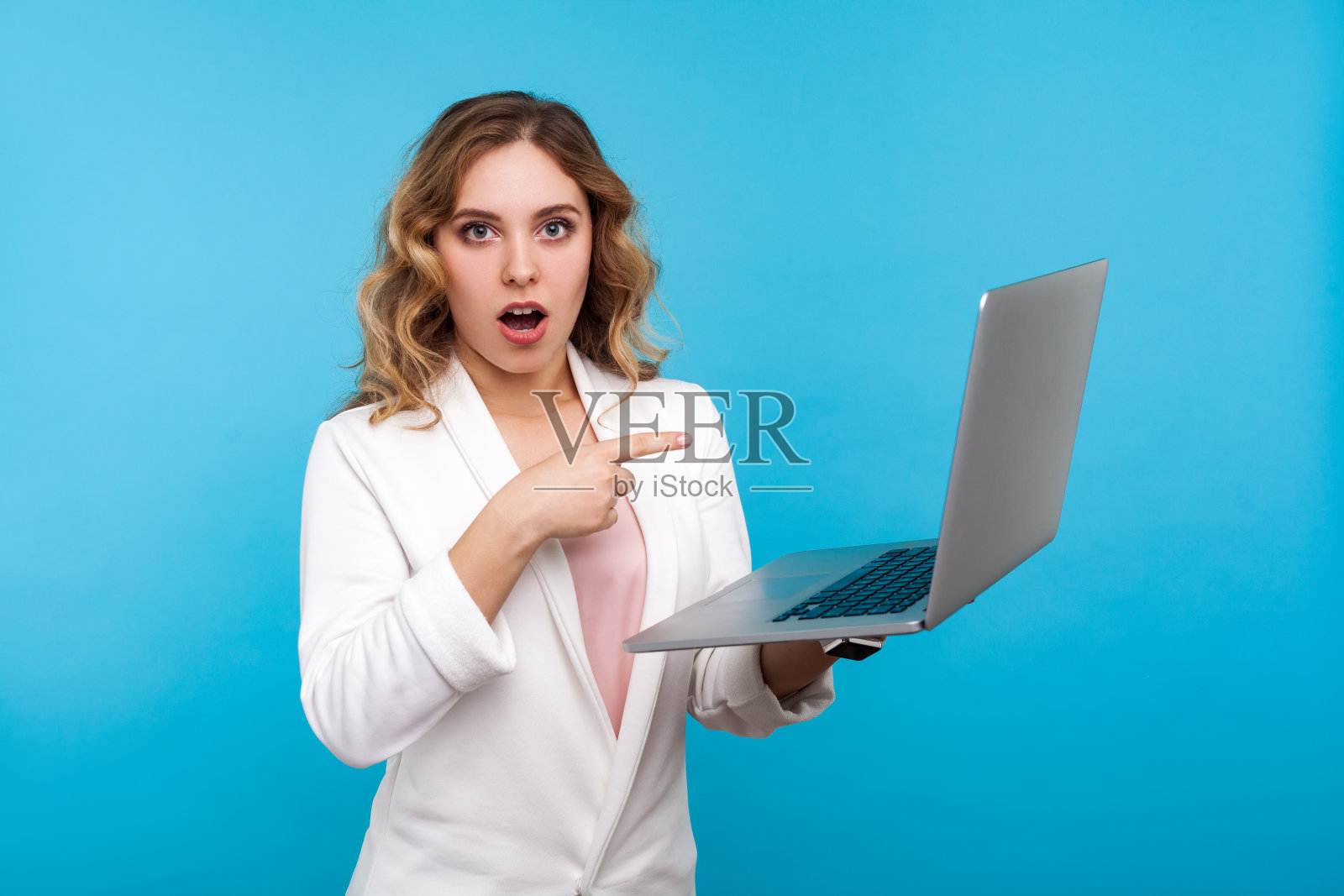 一名惊讶的女商人指着笔记本电脑，惊讶地看着相机。蓝色背景照片摄影图片