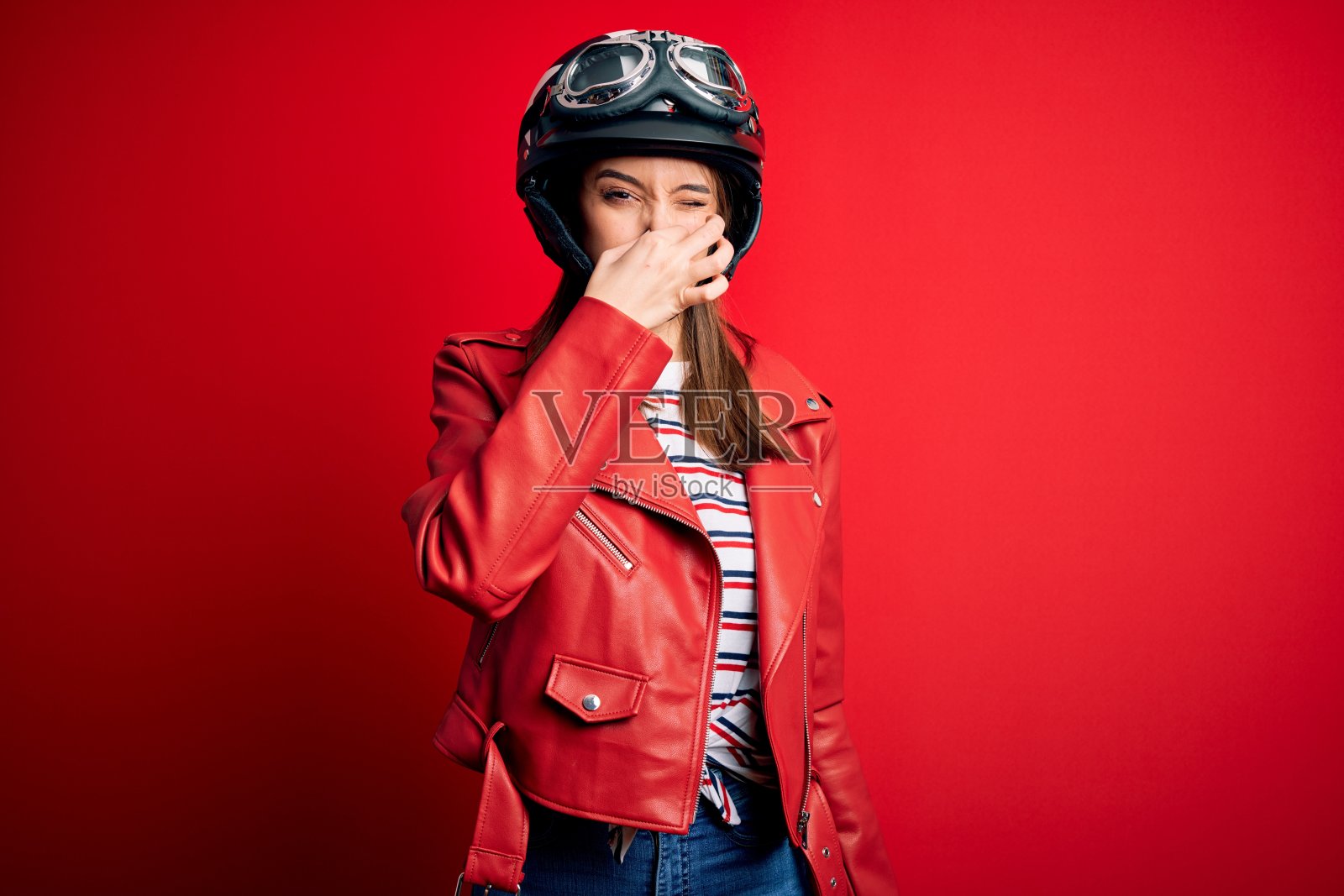 年轻漂亮的黑发摩托车女戴着摩托车头盔和红色夹克闻着一些臭气和恶心，难以忍受的气味，屏住呼吸用手指在鼻子上。不好的气味照片摄影图片