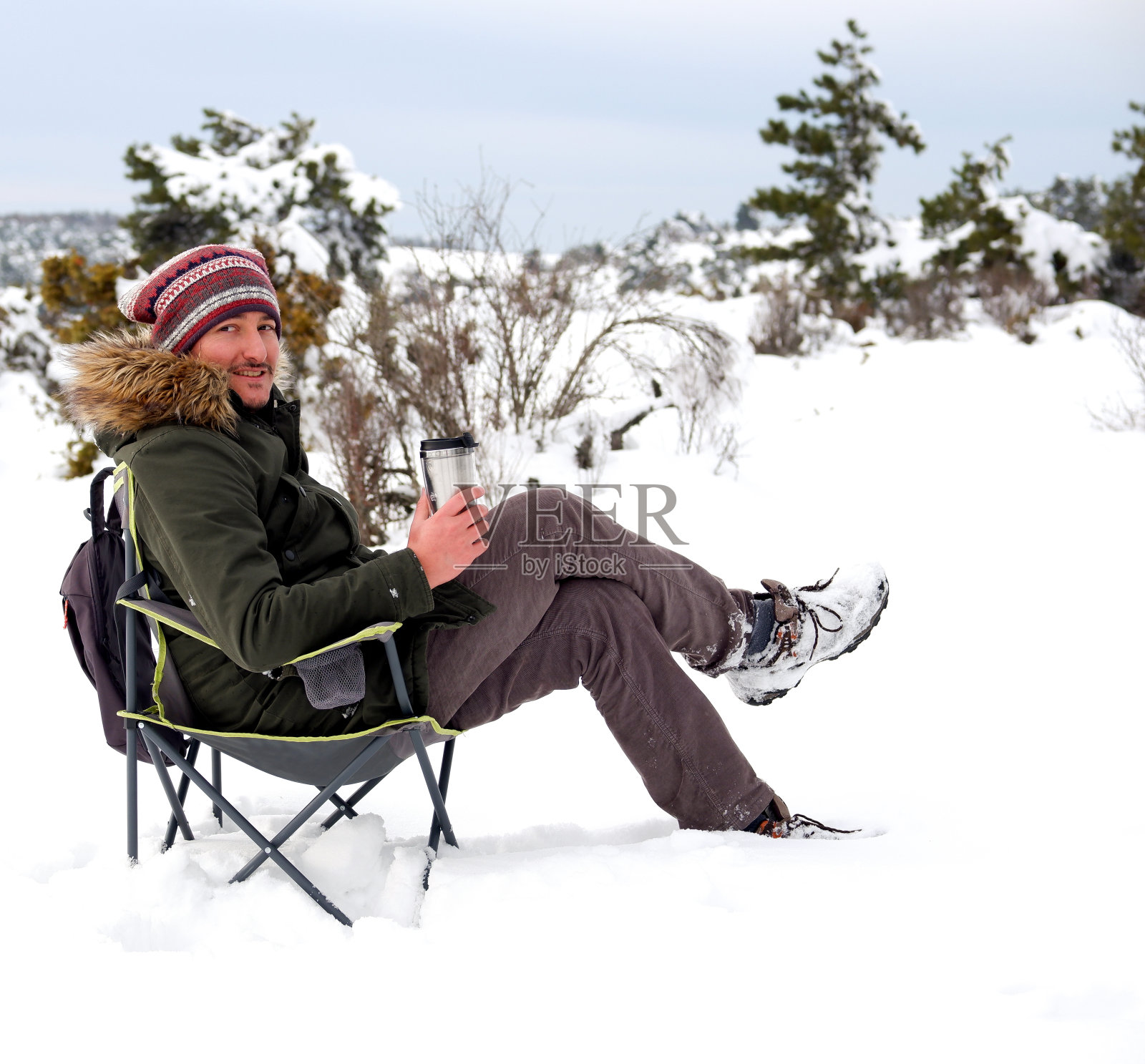 一个年轻人正坐在露营椅上，用保温杯喝着咖啡，享受着雪花照片摄影图片