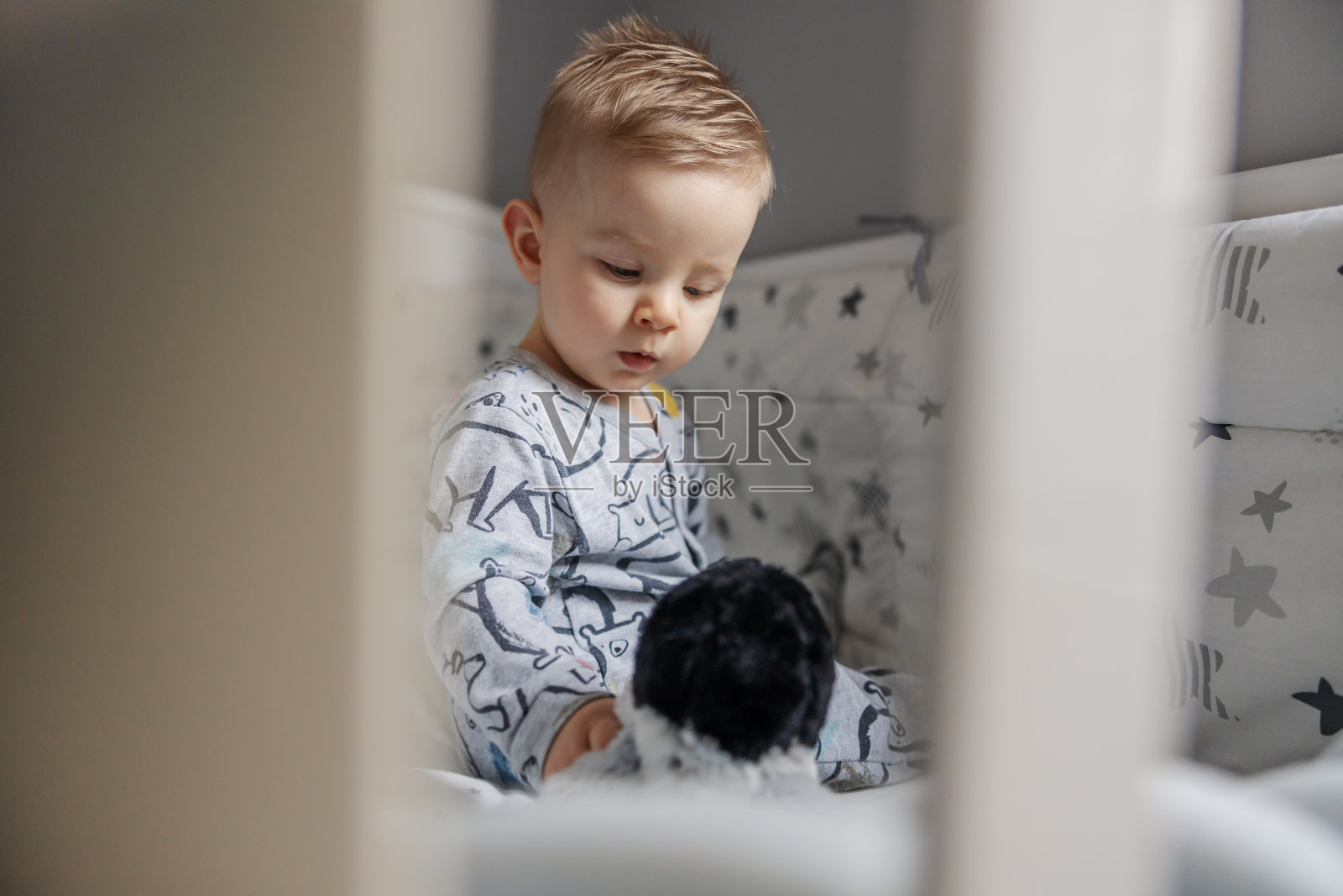 可爱的金发小男孩坐在他的婴儿床上，和玩具玩和说话的肖像。早上的时间。照片摄影图片