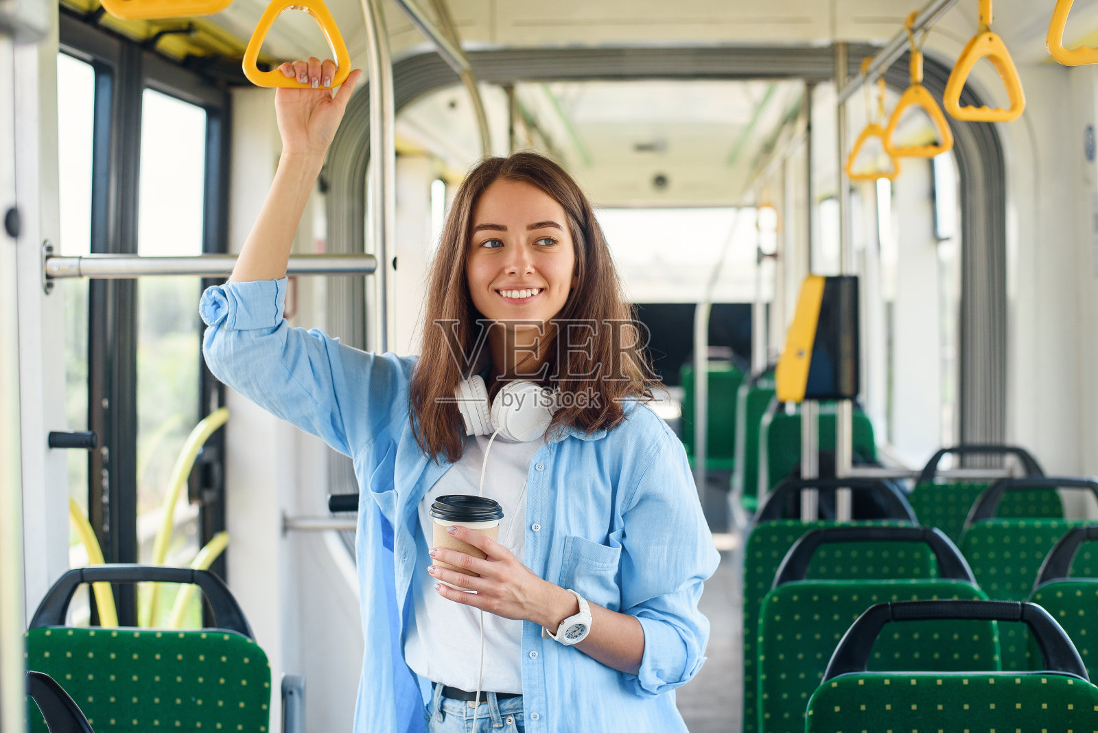 穿着蓝色衬衫的时尚女性在现代的有轨电车或公共汽车上享受旅行，在公共交通工具站着喝咖啡。照片摄影图片