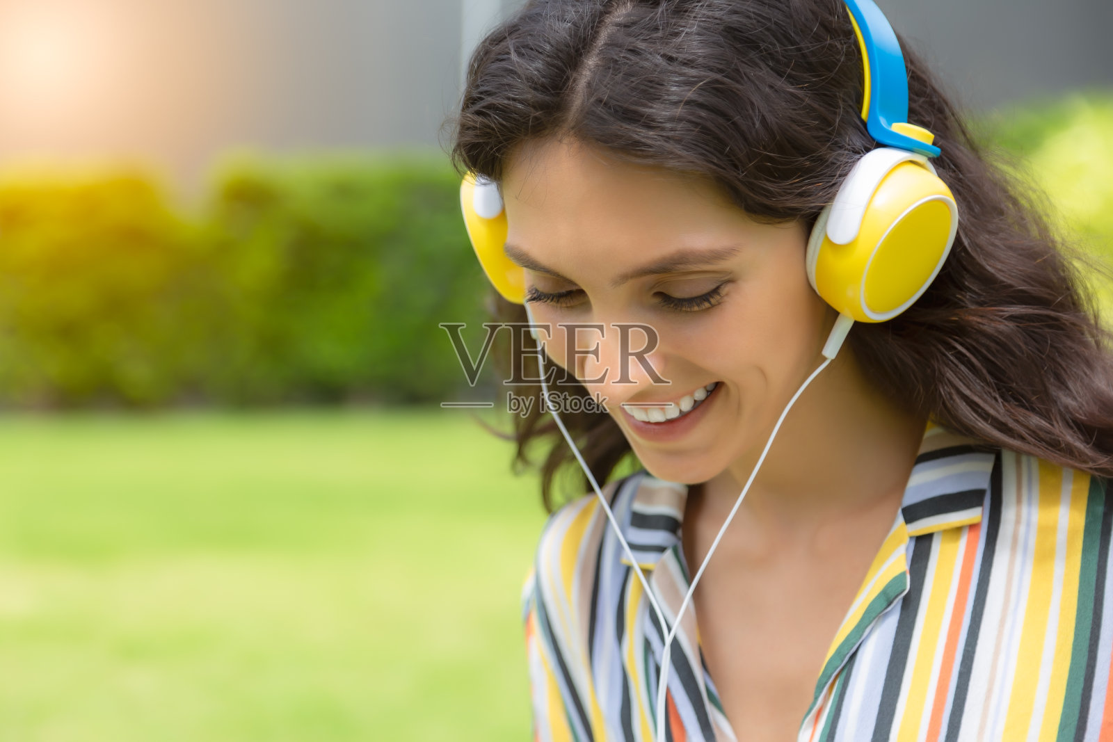 年轻女子用耳机听着美妙的音乐，脸上带着微笑，在夏天的公园里快乐。美女放松，享受生活。漂亮女人喜欢夏天。她喜欢听音乐照片摄影图片