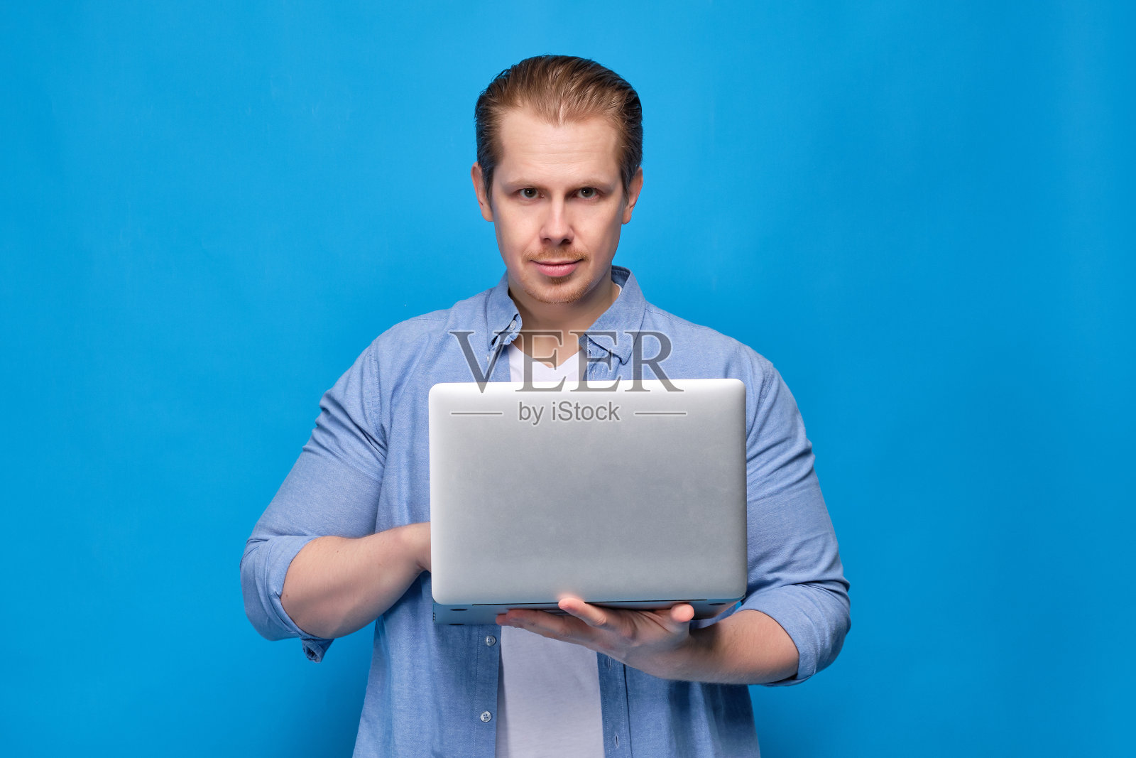 一名身穿蓝色衬衫的男子拿着一台笔记本电脑，看着摄像头。处理故障排除的程序员概念。照片摄影图片