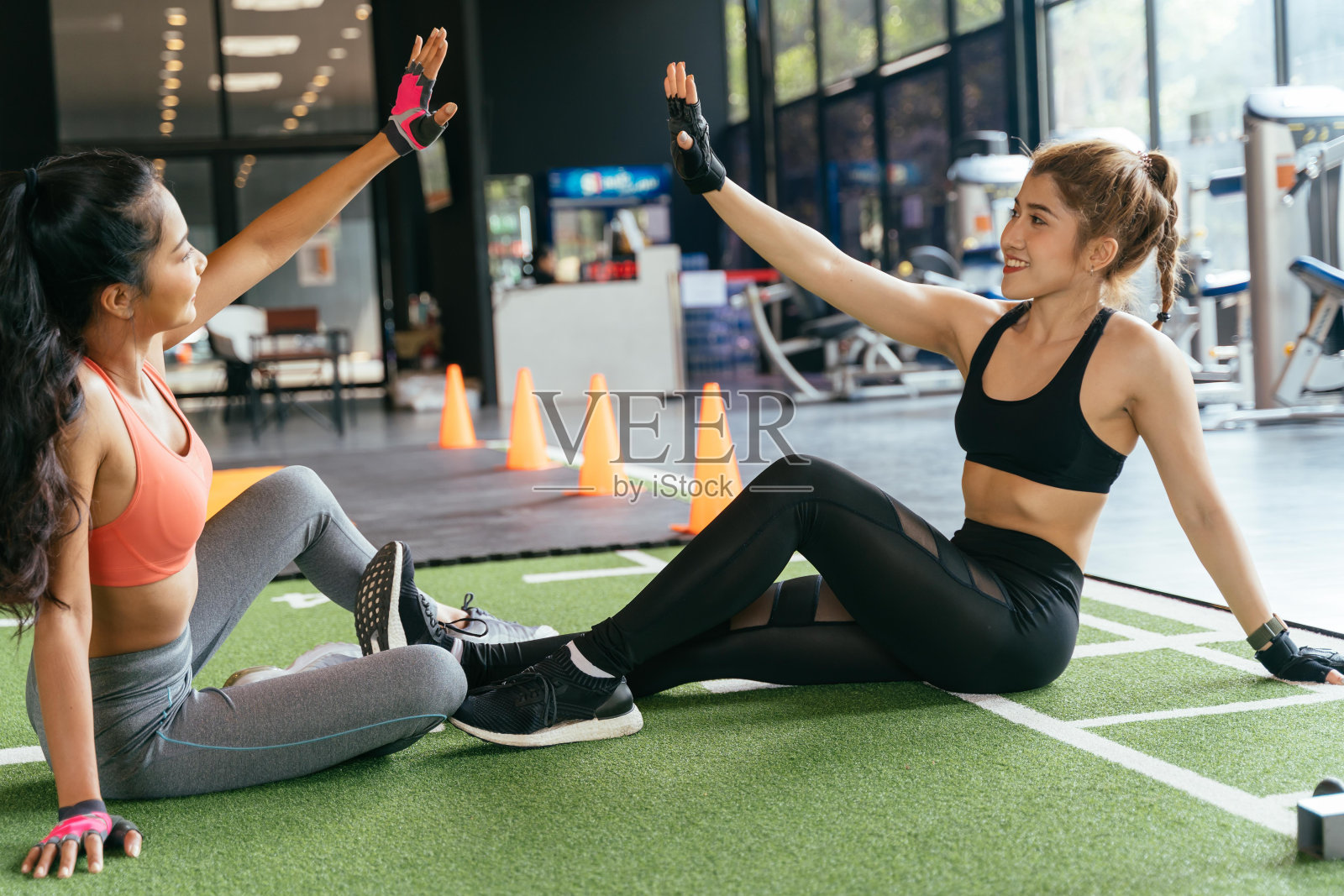 健康微笑的女性朋友们坐在健身房的地板上互相击掌。亚洲的运动人士一起锻炼。照片摄影图片