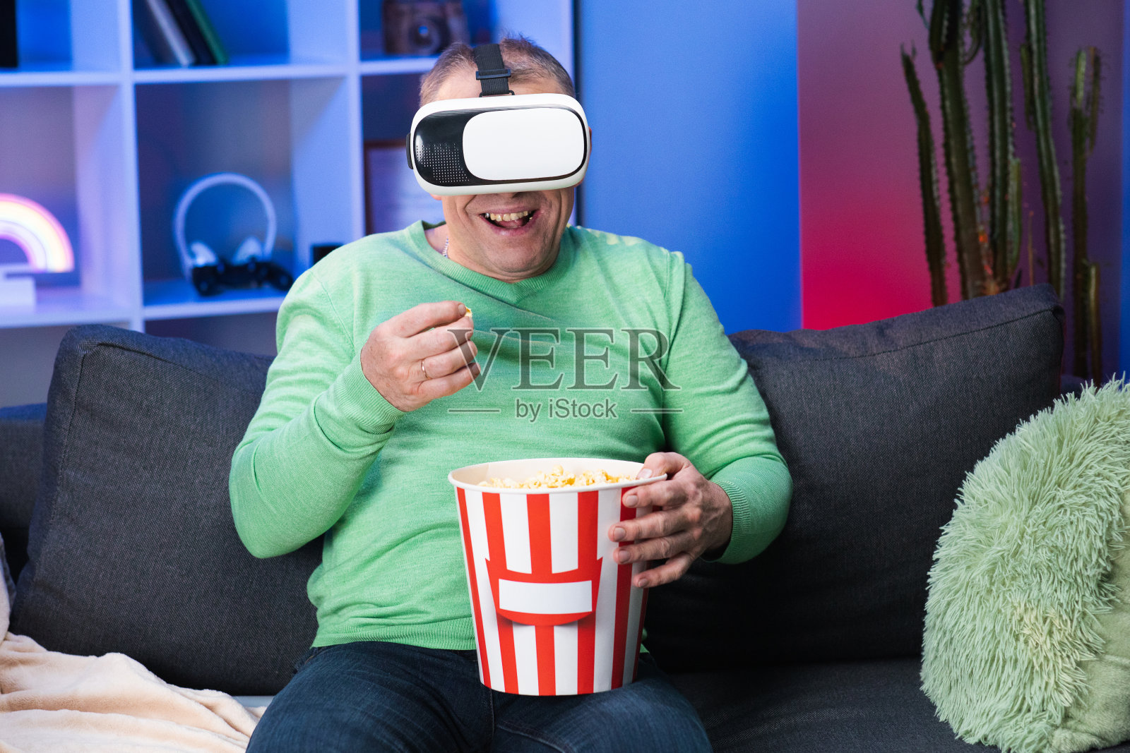 白人男子与一盒爆米花在他的手看视频使用虚拟现实耳机坐在沙发上吃爆米花在房间照片摄影图片