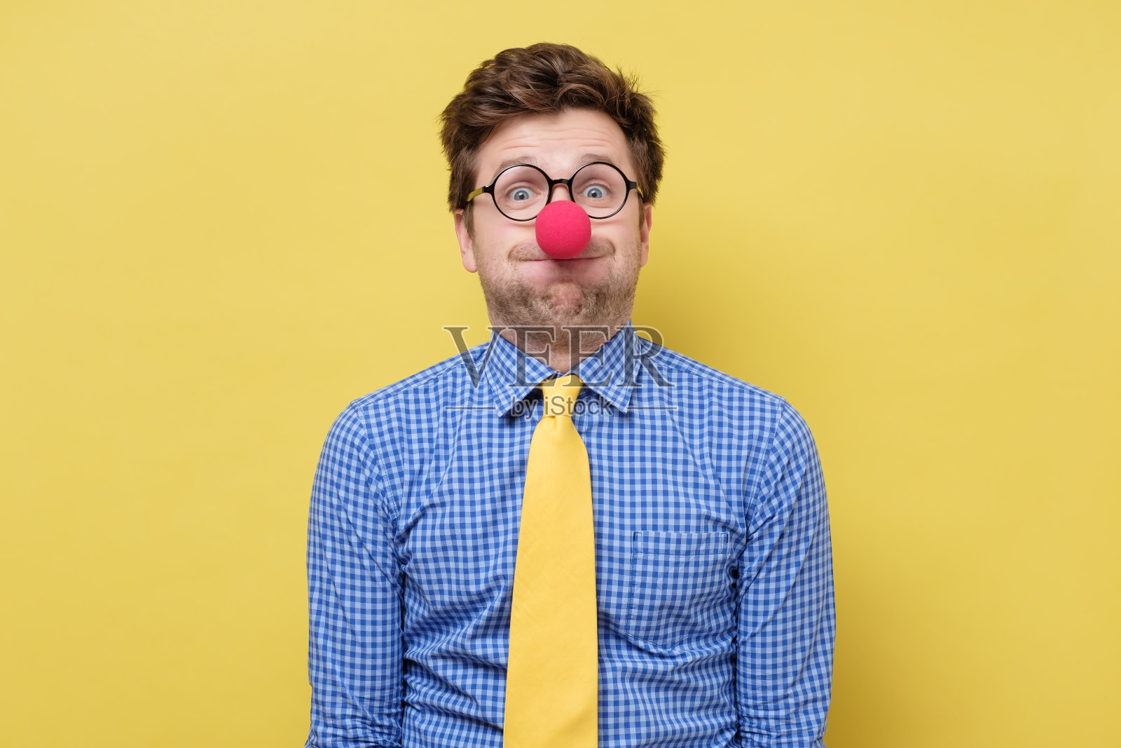 年轻英俊的男人用红色的小丑鼻子吹着脸颊。照片摄影图片