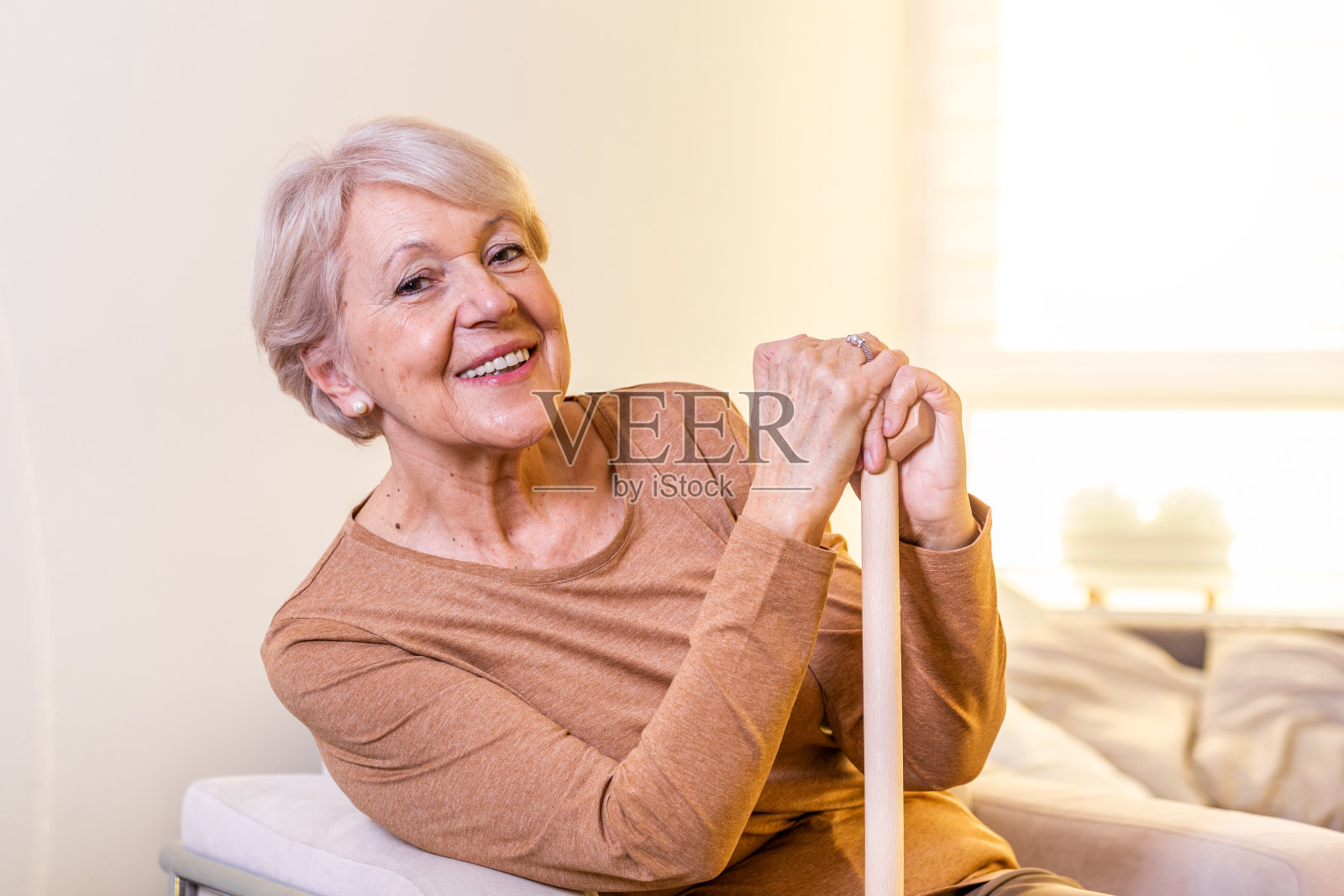 老妇人坐在沙发上，拄着木拐杖，面带微笑。快乐的老妇人在沙发上放松，拿着手杖。副本的空间。一位看起来很体贴的老妇人在养老院照片摄影图片