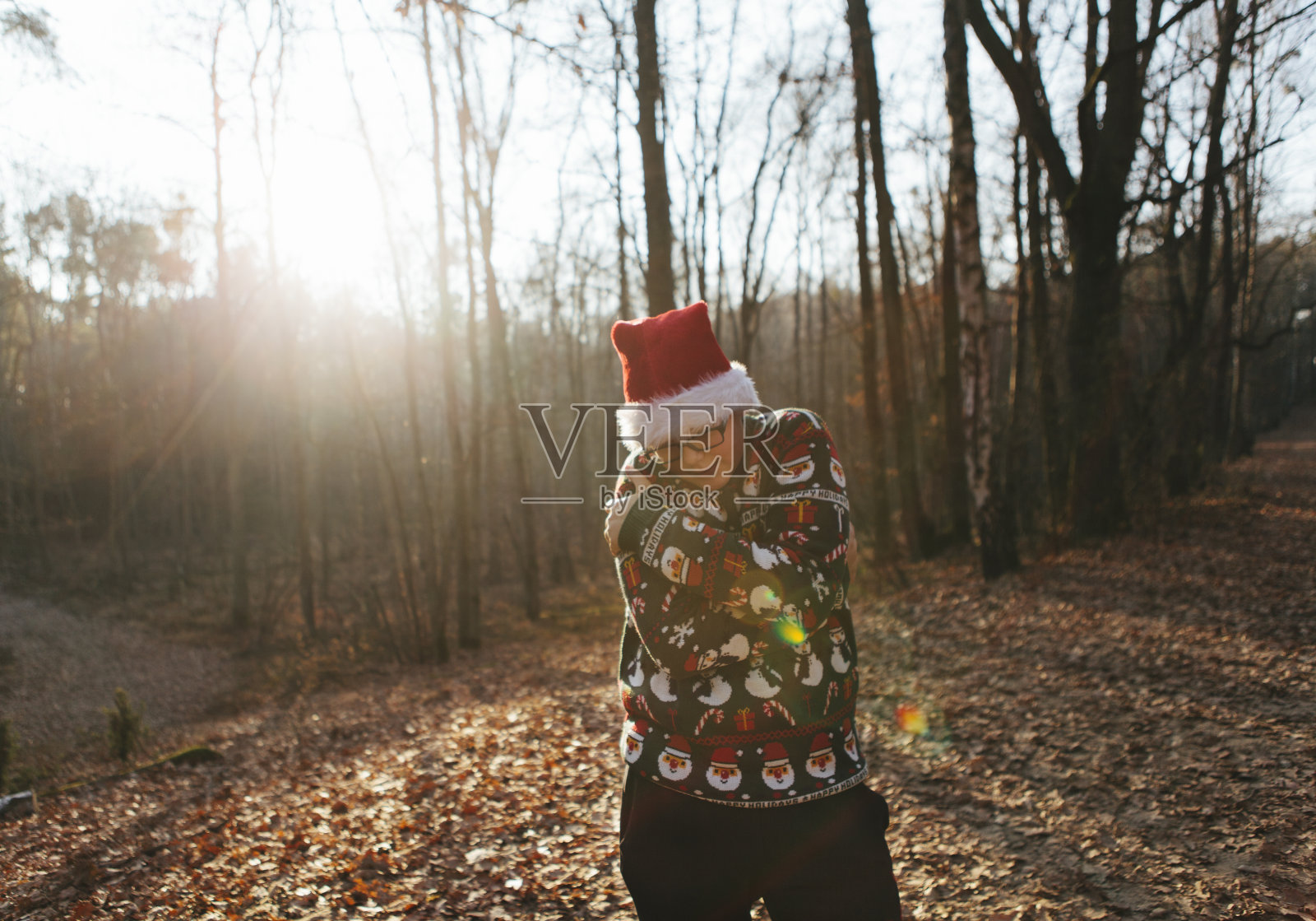 穿着圣诞毛衣，戴着圣诞老人帽的帅气少年独自站在冬日的阳光下，在寒冷中拥抱自己照片摄影图片