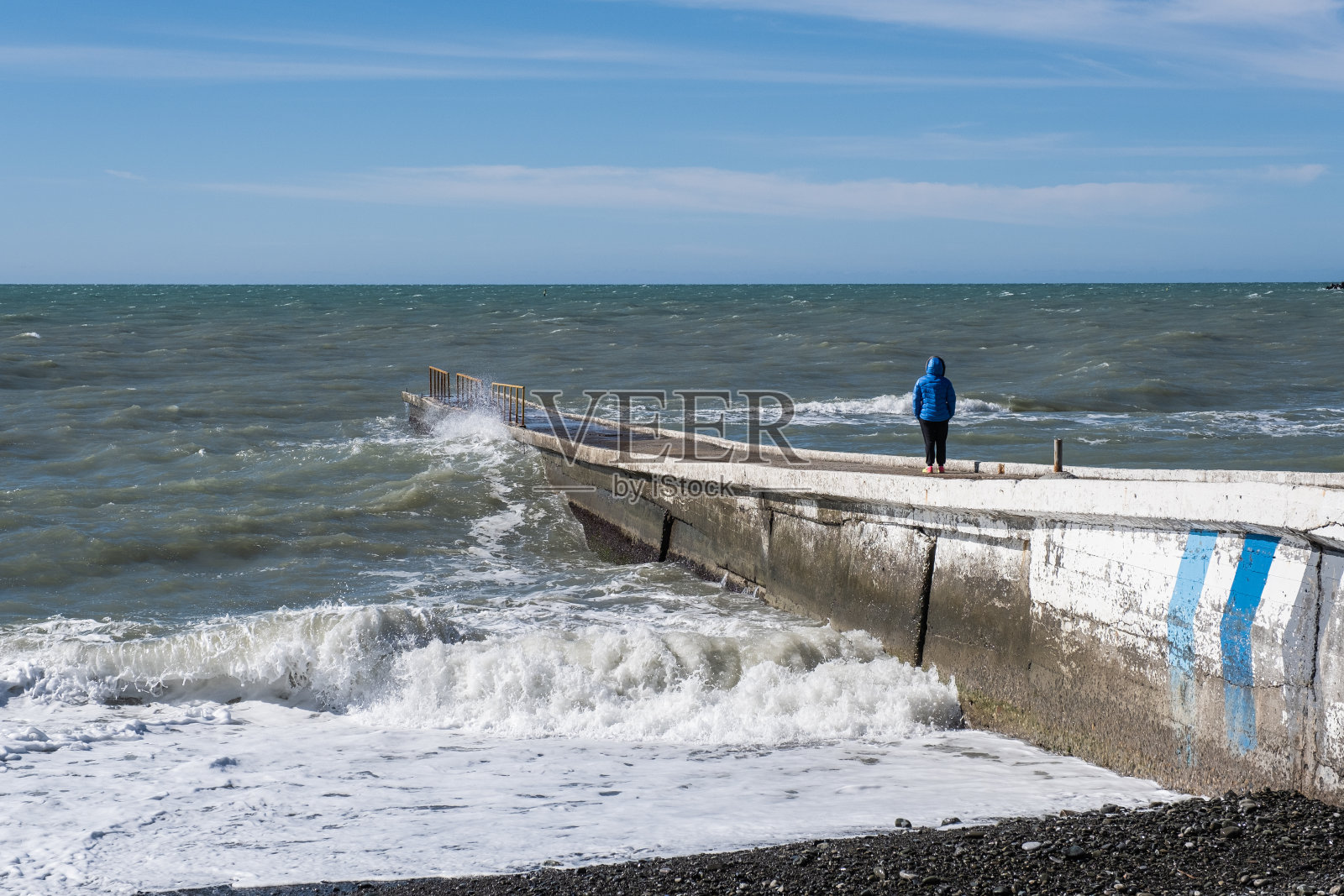 冬天的海景观。一个穿着蓝色夹克的女孩站在防波堤上看着海浪。照片摄影图片