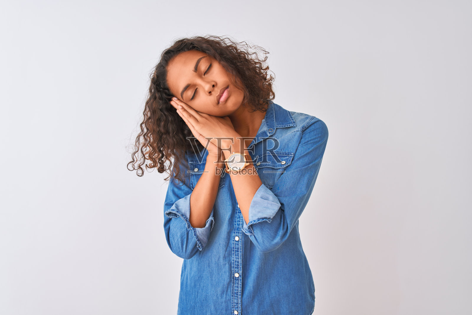 年轻的巴西女子穿着牛仔衬衫站在孤立的白色背景上，疲倦地睡觉，做着梦，闭着眼睛微笑着双手合在一起。照片摄影图片