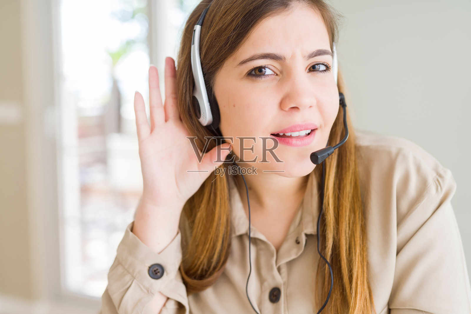 年轻漂亮的女接线员戴着耳机在办公室微笑，手捂着耳朵听谣言或流言蜚语。耳聋的概念。照片摄影图片