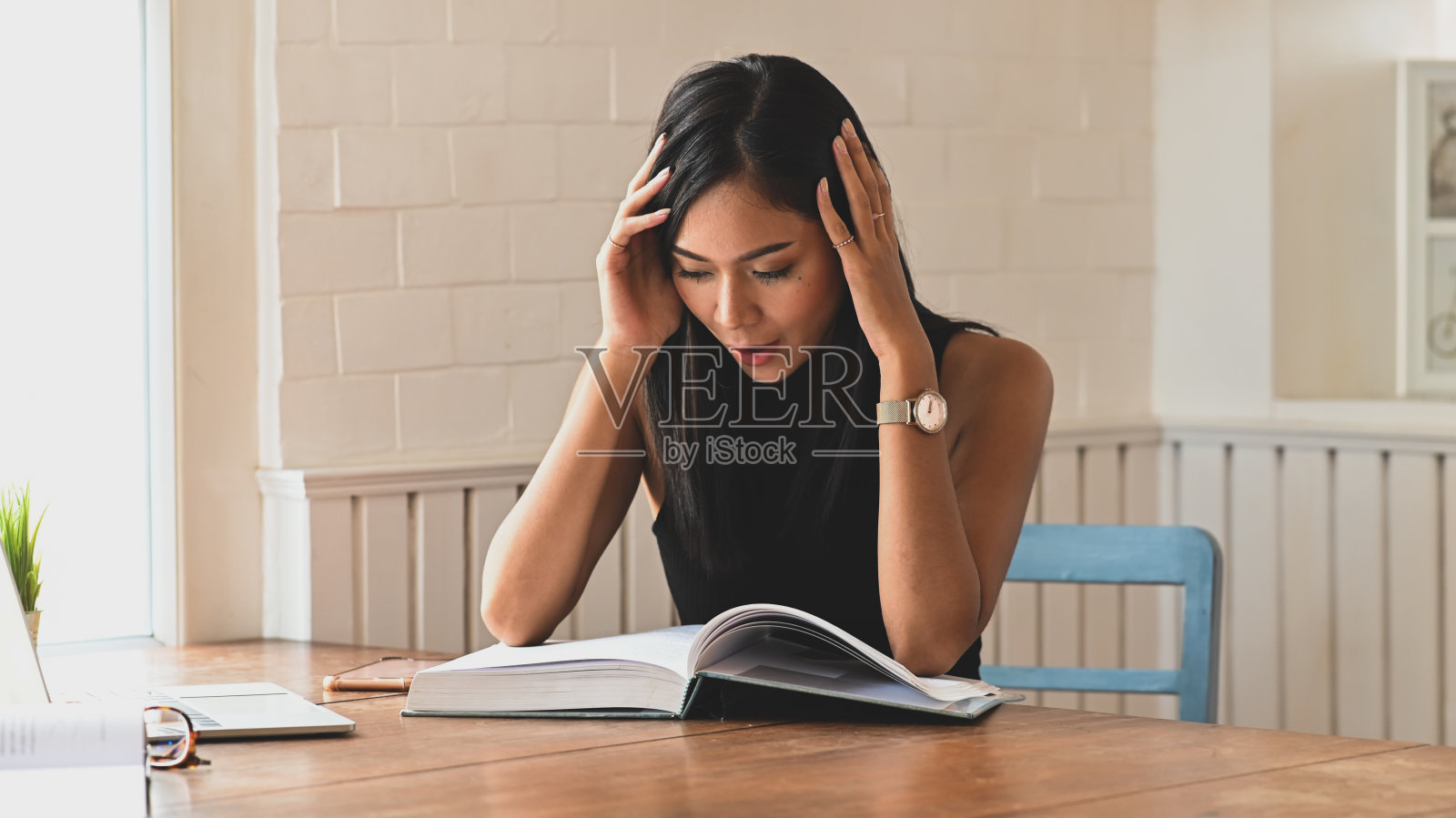 坐在以舒适客厅为背景的木桌前看书时，手放在头上的压力大的女人。照片摄影图片