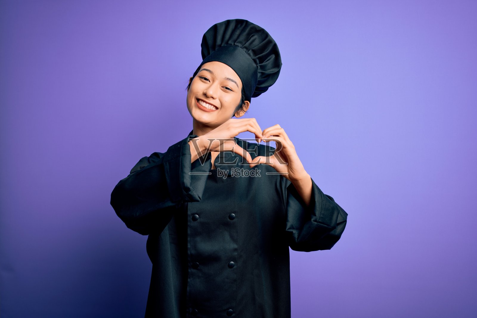 年轻美丽的中国女厨师穿着炊具制服和帽子在紫色的背景下微笑着做心形符号形状的手。浪漫的概念。照片摄影图片