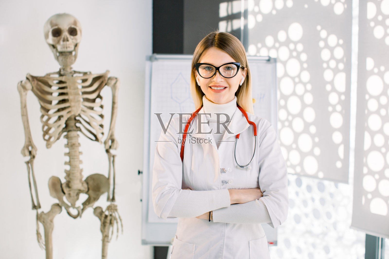 漂亮的白人女医生的肖像听诊器，穿着白大褂和眼镜，微笑和交叉双臂在胸前，站在现代的办公室。人类骨骼和剪贴板的背景照片摄影图片