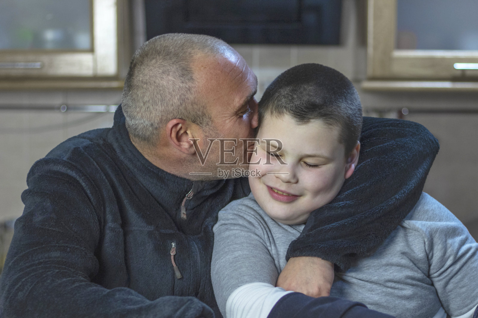 自闭症的爸爸和他的儿子坐在家里的厨房里。一个留着短头发、鬃毛的男人拥抱他的儿子，亲吻他的脸颊。照片摄影图片