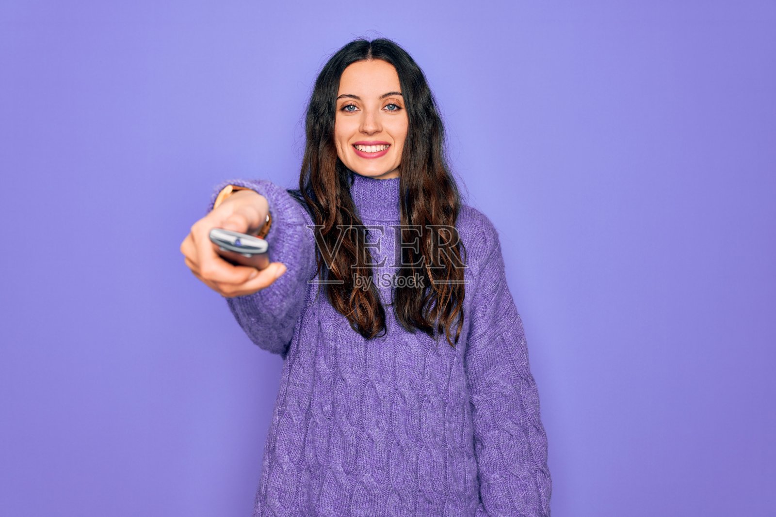 年轻美丽的女人用电视遥控器控制孤立的紫色背景，一个快乐的脸站着，微笑着自信的微笑显示牙齿照片摄影图片