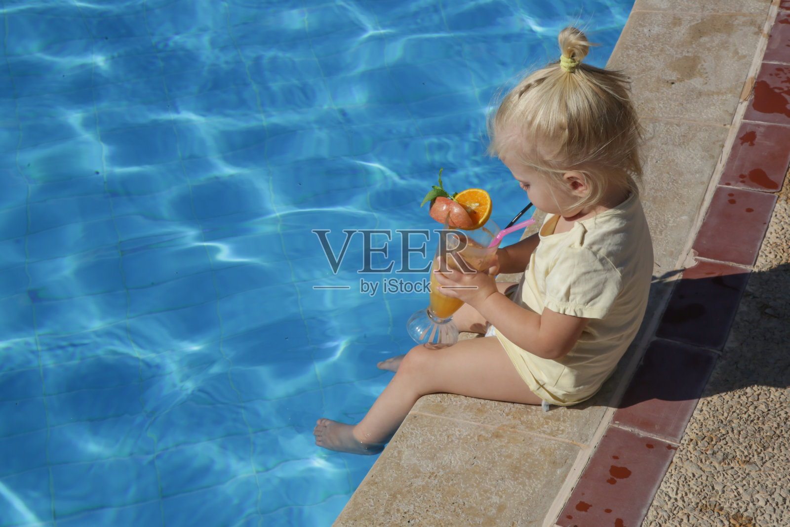 可爱的金发小女孩坐在泳池边，喝着新鲜的橙汁，在炎热的夏天在游泳池里度过一天，补充水分和维生素照片摄影图片