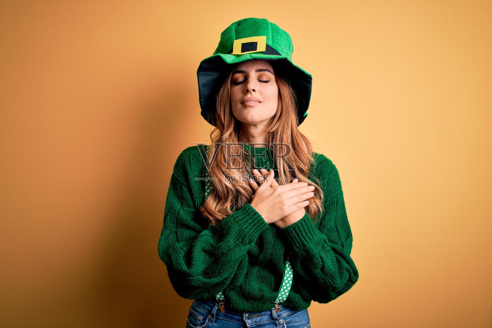 美丽的黑发女人戴着绿帽子与三叶草庆祝圣帕特里克节微笑与手在胸前闭上眼睛和感激的手势在脸上。健康的概念。照片摄影图片