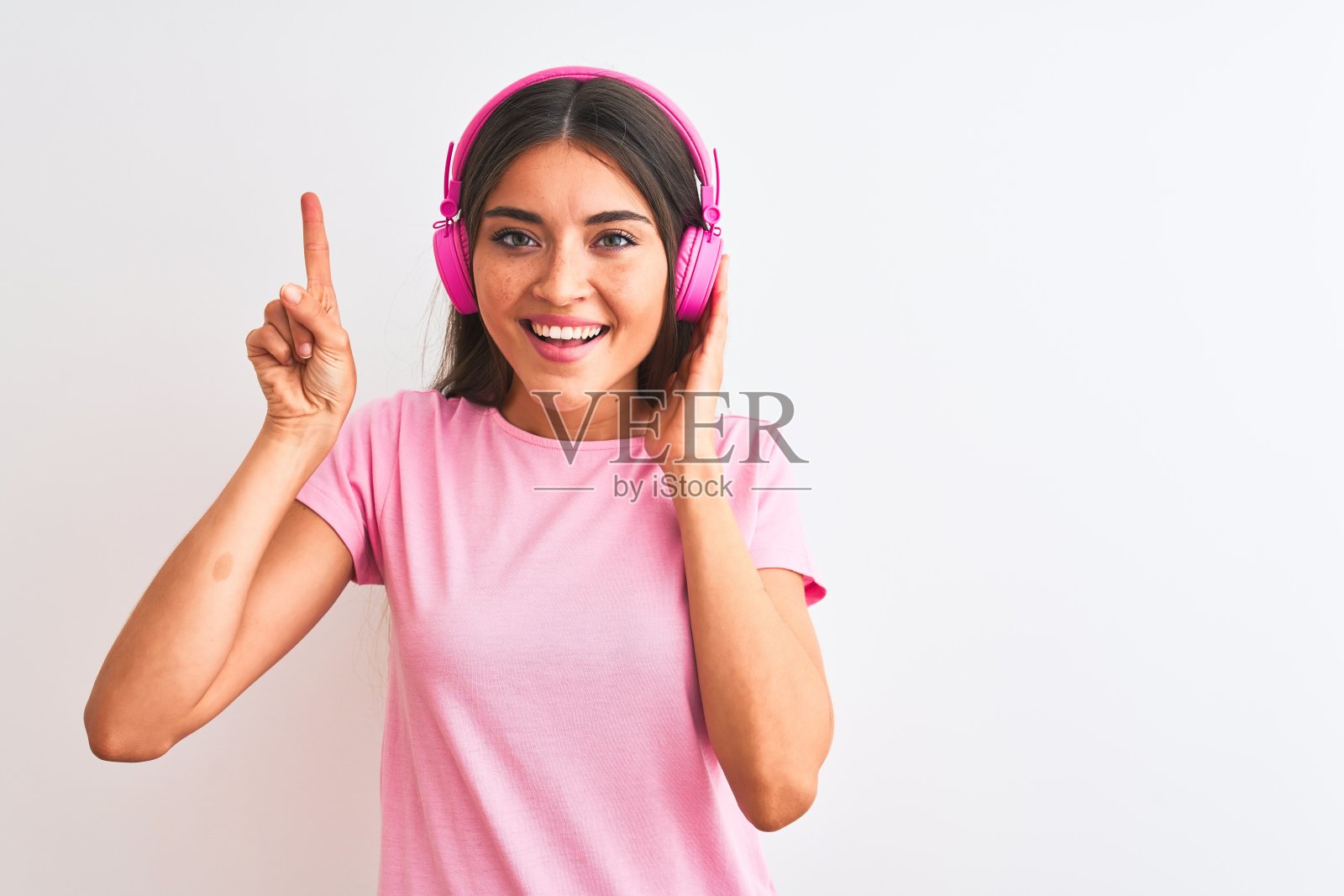 年轻漂亮的女人在孤立的白色背景下戴着耳机听音乐，惊讶于一个想法或问题，开心地指着手指，第一照片摄影图片