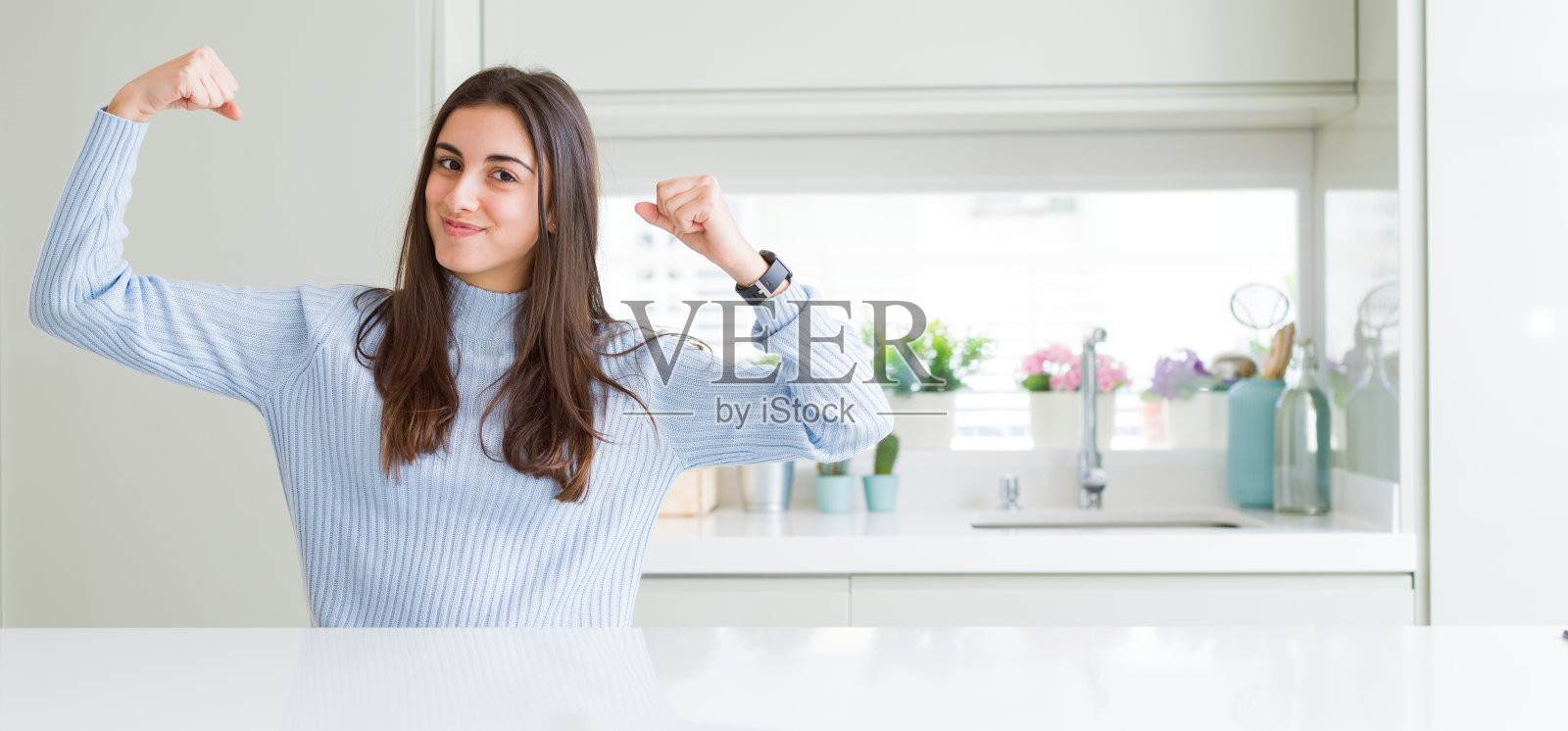 广角图片美丽的年轻女子坐在白色的桌子在家里显示手臂肌肉微笑自豪。健康的概念。照片摄影图片