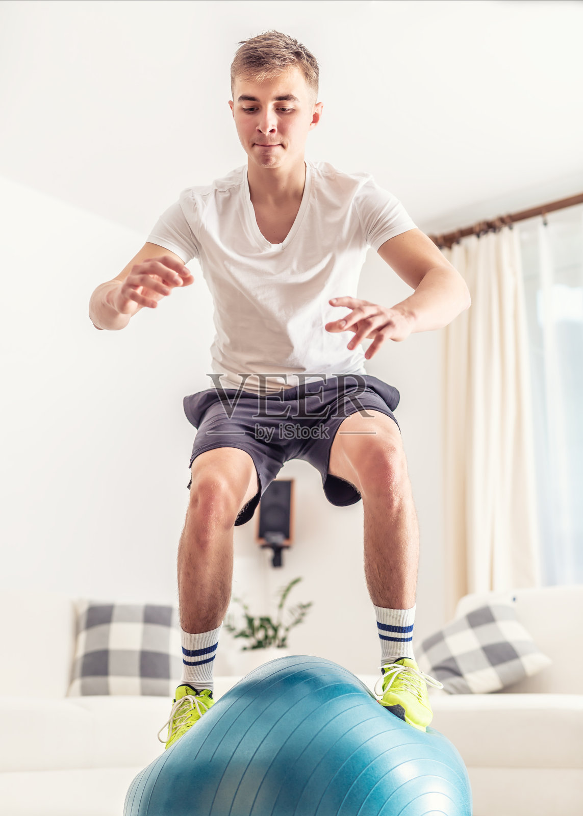在斯堪的那维亚风格的公寓里，运动的年轻男性在家庭训练中，穿着t恤，短裤和运动鞋完成一个蓝色的适合球。照片摄影图片