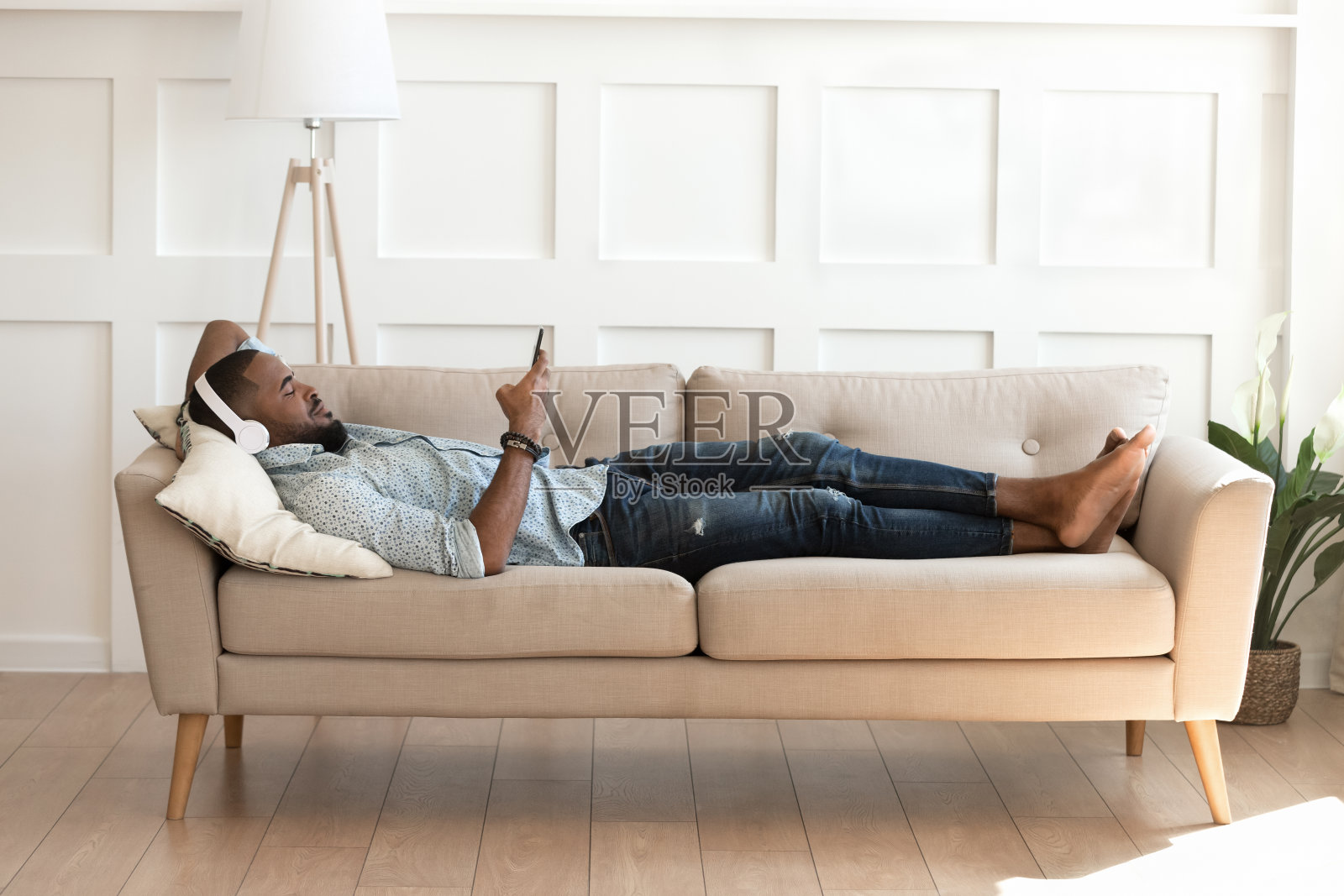 戴着耳机躺在沙发上看智能手机的非洲男人照片摄影图片