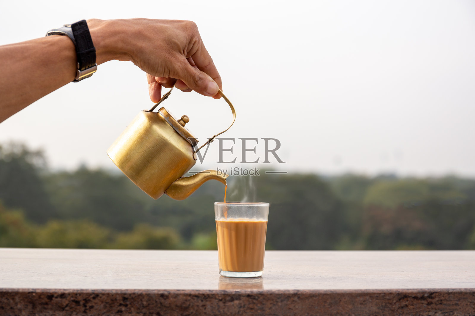 用手将马沙拉茶从茶壶倒入玻璃杯中。照片摄影图片