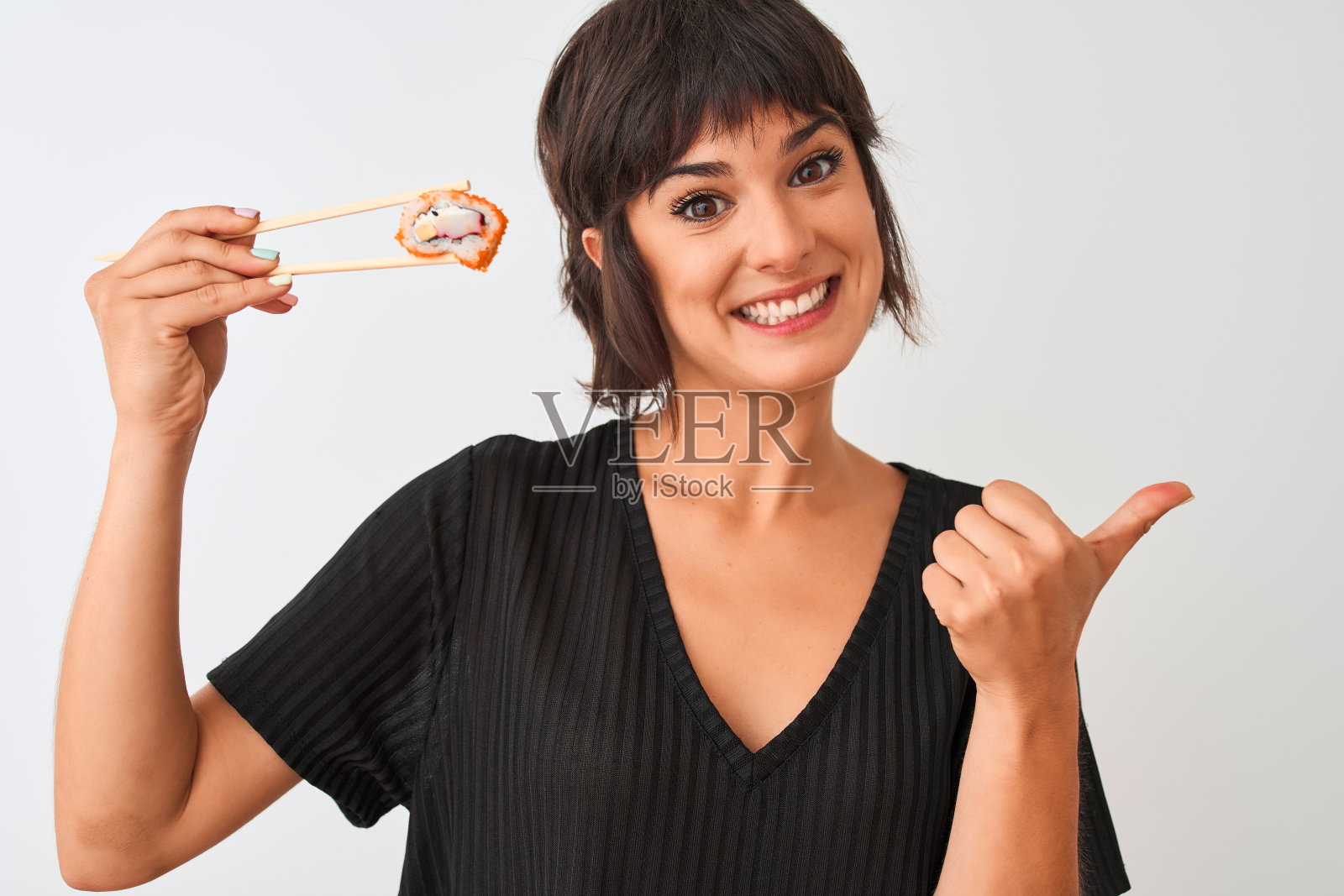 年轻美丽的女人吃寿司用筷子站在孤立的白色背景指向和显示拇指向上的一边与幸福的脸微笑照片摄影图片