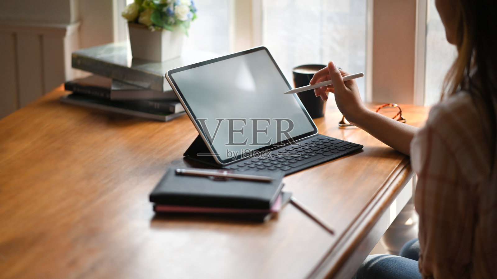 裁剪的形象，创造性的女人拿着一支笔，而绘图/指向白色空白屏幕的电脑平板电脑与键盘的情况下，把放在办公桌上舒适的房间作为背景。照片摄影图片