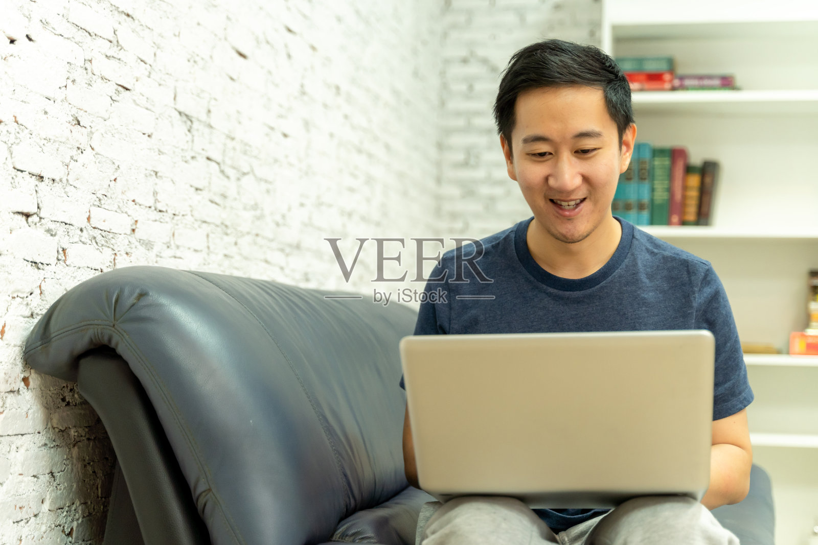 年轻的亚洲人在休闲服装在沙发上工作的笔记本电脑。男性自由职业者在家用笔记本工作。照片摄影图片
