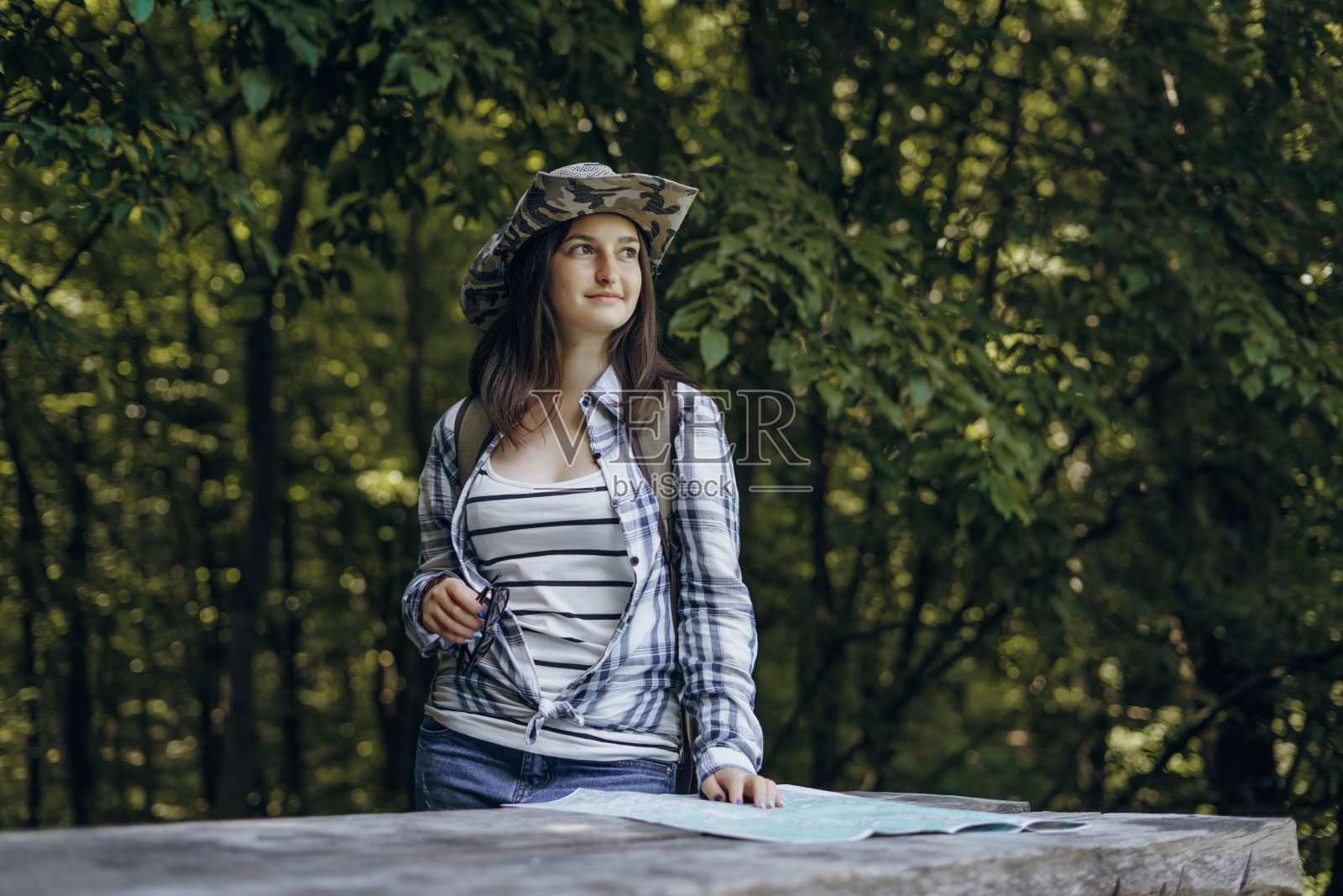 戴着帽子的黑发女孩在夏天的森林里带着旅游地图。照片摄影图片