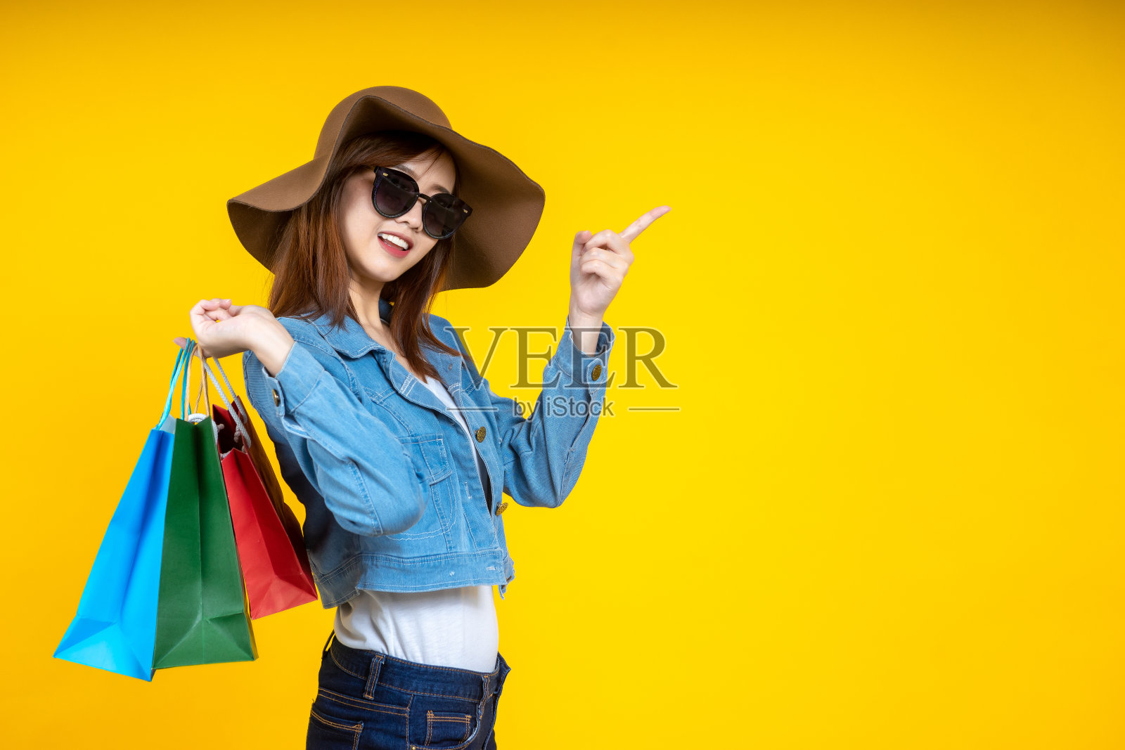 亚洲女人漂亮迷人的肖像戴太阳眼镜携带新的购物袋点手指复制空间上明亮孤立的黄色背景。购物的概念。照片摄影图片