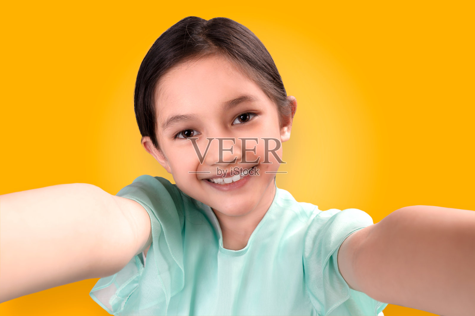 漂亮的亚洲孩子在自拍，黄色的背景上带着大大的微笑，很有趣。可爱的孩子横伸双手拿着相机自拍或用智能手机拍视频照片摄影图片
