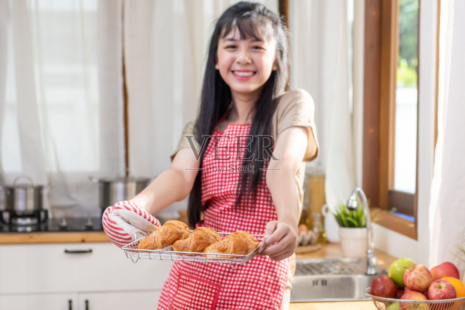 微笑年轻美丽的亚洲女人女孩与围裙拿着一盘新鲜美味的羊角面包在家庭厨房。迷人的少女享受和有乐趣学习烘焙和烹饪课在家里。照片摄影图片