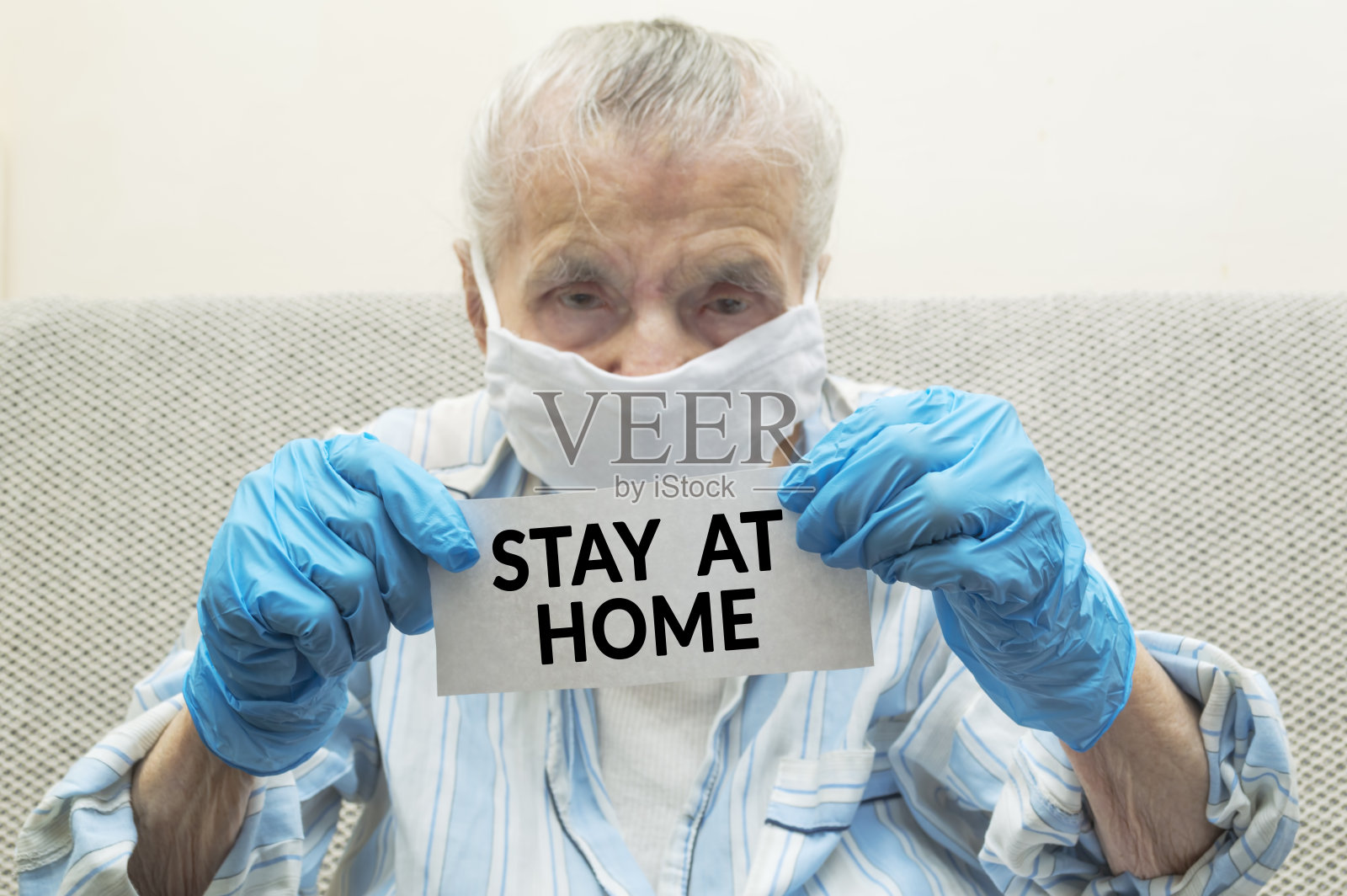 一位脸上戴着医用防护口罩和防护手套的老年妇女手持写有“待在家里”字样的纸。照片摄影图片