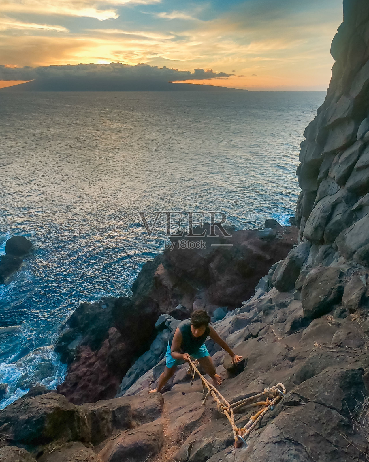 一名男子在爬下夏威夷毛伊岛海岸悬崖边缘的岩壁时找到了一个立足点。无尽的海洋。攀爬者的腿。绳子没有固定。宽镜头。照片摄影图片