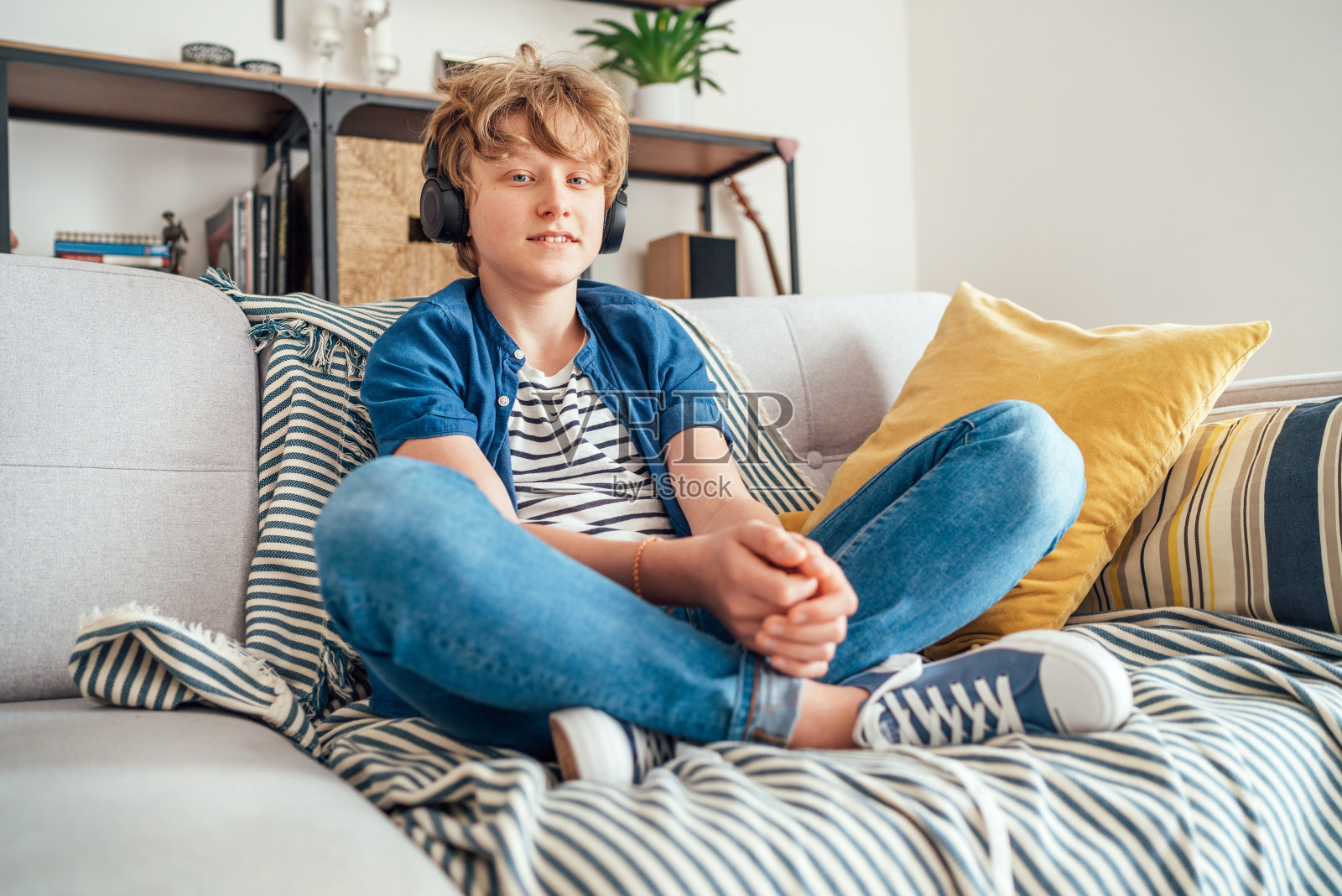 一个十几岁的小男孩盘腿坐在家里舒适的沙发上，穿着休闲牛仔裤和新运动鞋，戴着无线耳机听着音乐，对着镜头微笑照片摄影图片