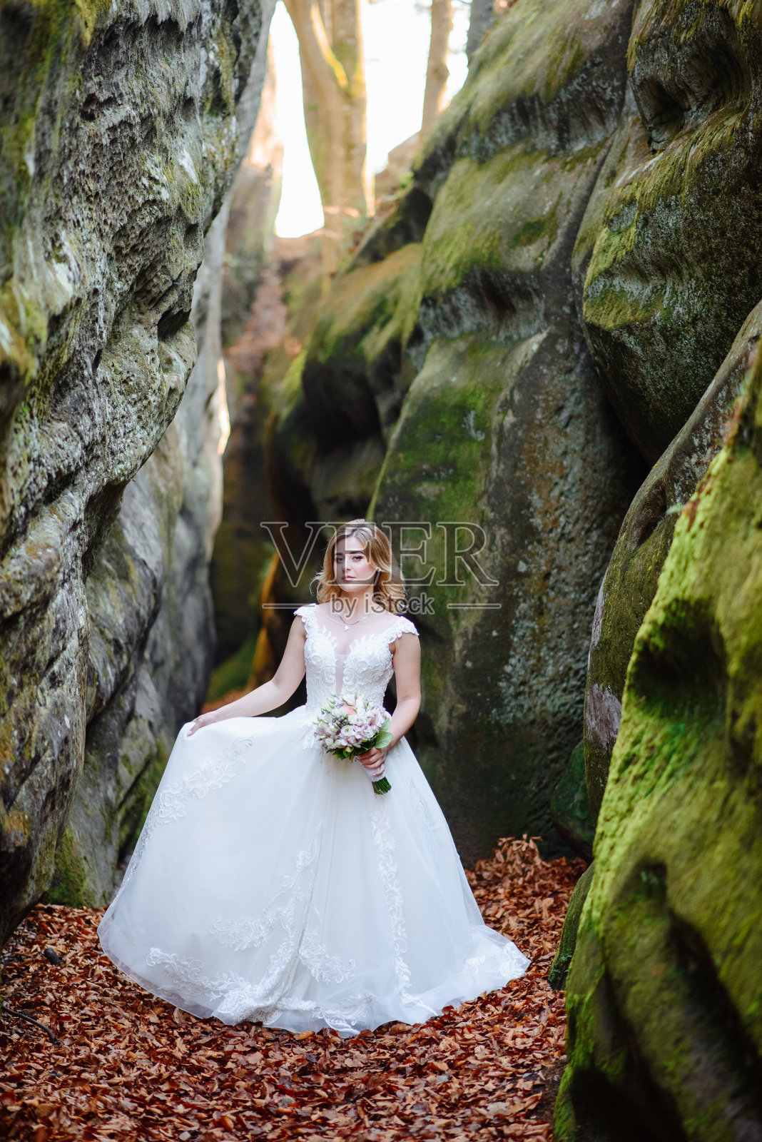 美丽的金发新娘捧着一束鲜花站在狭窄的峡谷里。照片摄影图片