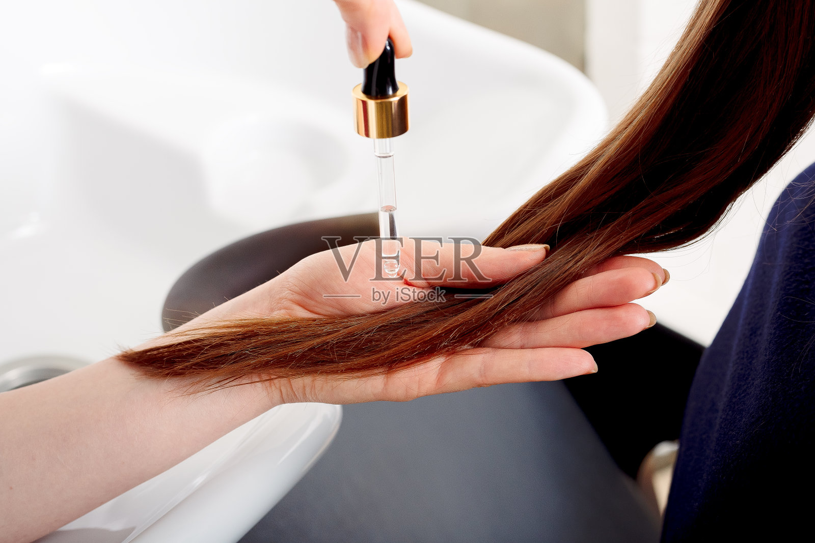 女性的手在长长的棕色头发上涂油。护发化妆品、沐浴美容spa产品。女性护发精华用于护理发梢对抗美容院室内、浴室照片摄影图片