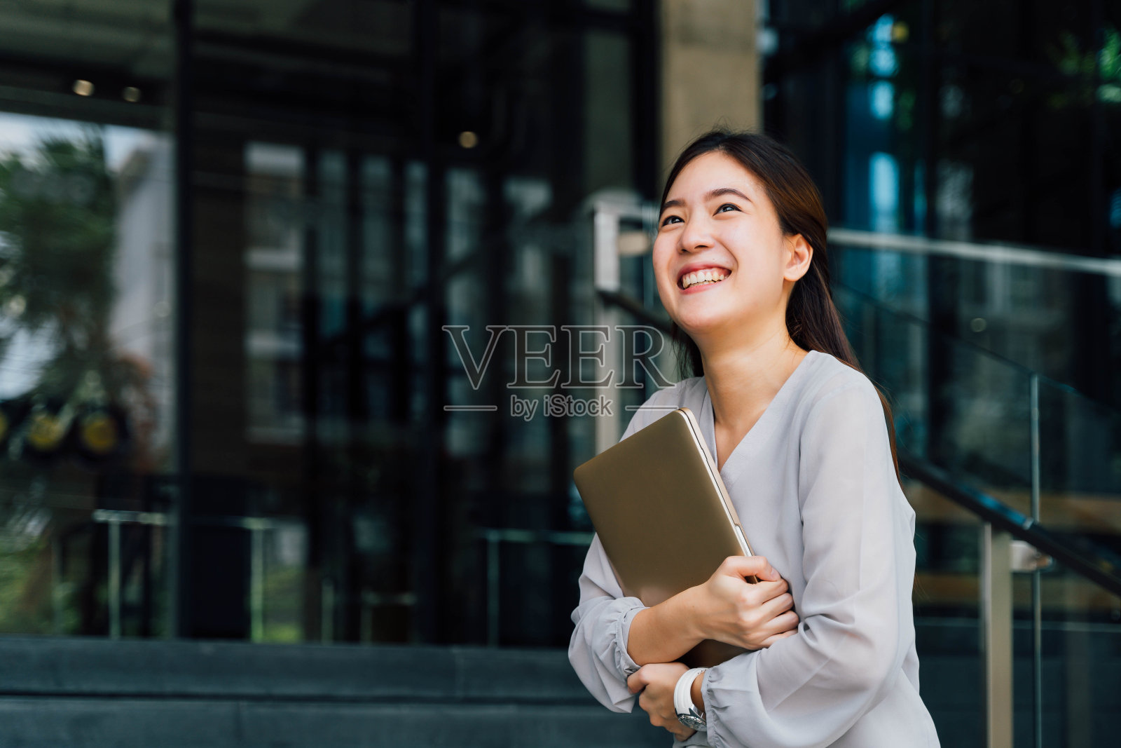 一位年轻成功的商业女性微笑着拿着一台笔记本电脑。职业女性工作者站在公司现代办公室和窗户在休闲灰色的样子。-有拷贝空间照片摄影图片