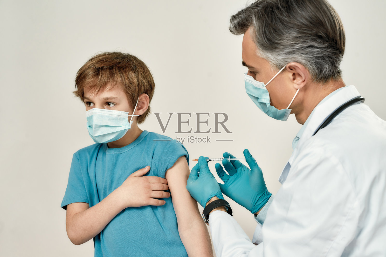 接受疫苗。专业成熟的医生以医用口罩和蓝色无菌手套为小男孩打针。免疫、医药和保健照片摄影图片