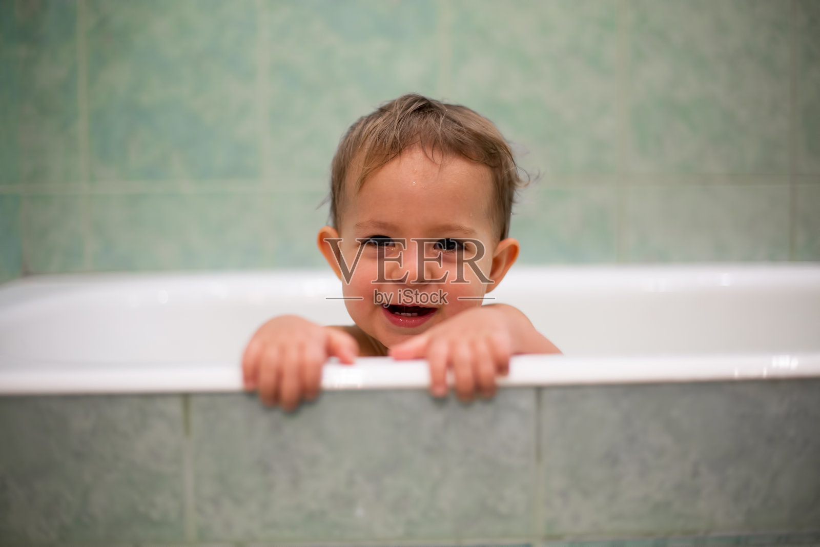 一个可爱的白人婴儿从浴缸里探出头来，把手放在浴缸边，看着镜头，张大嘴巴笑了。背景是一个模糊的绿色浴室。特写,软焦点照片摄影图片