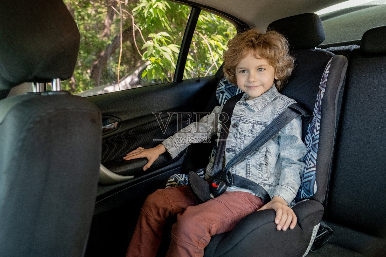 快乐可爱的小男孩在牛仔夹克和棕色裤子坐在车里照片摄影图片