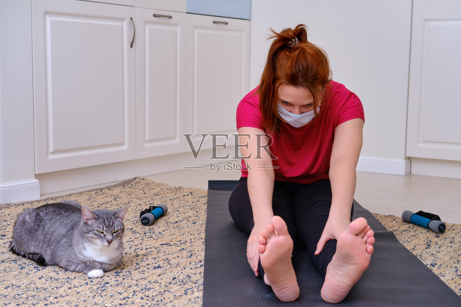 一个带着医用口罩的女人和一只猫在家庭厨房里做瑜伽。由于冠状病毒大流行，隔离期间有独立的健身课程照片摄影图片