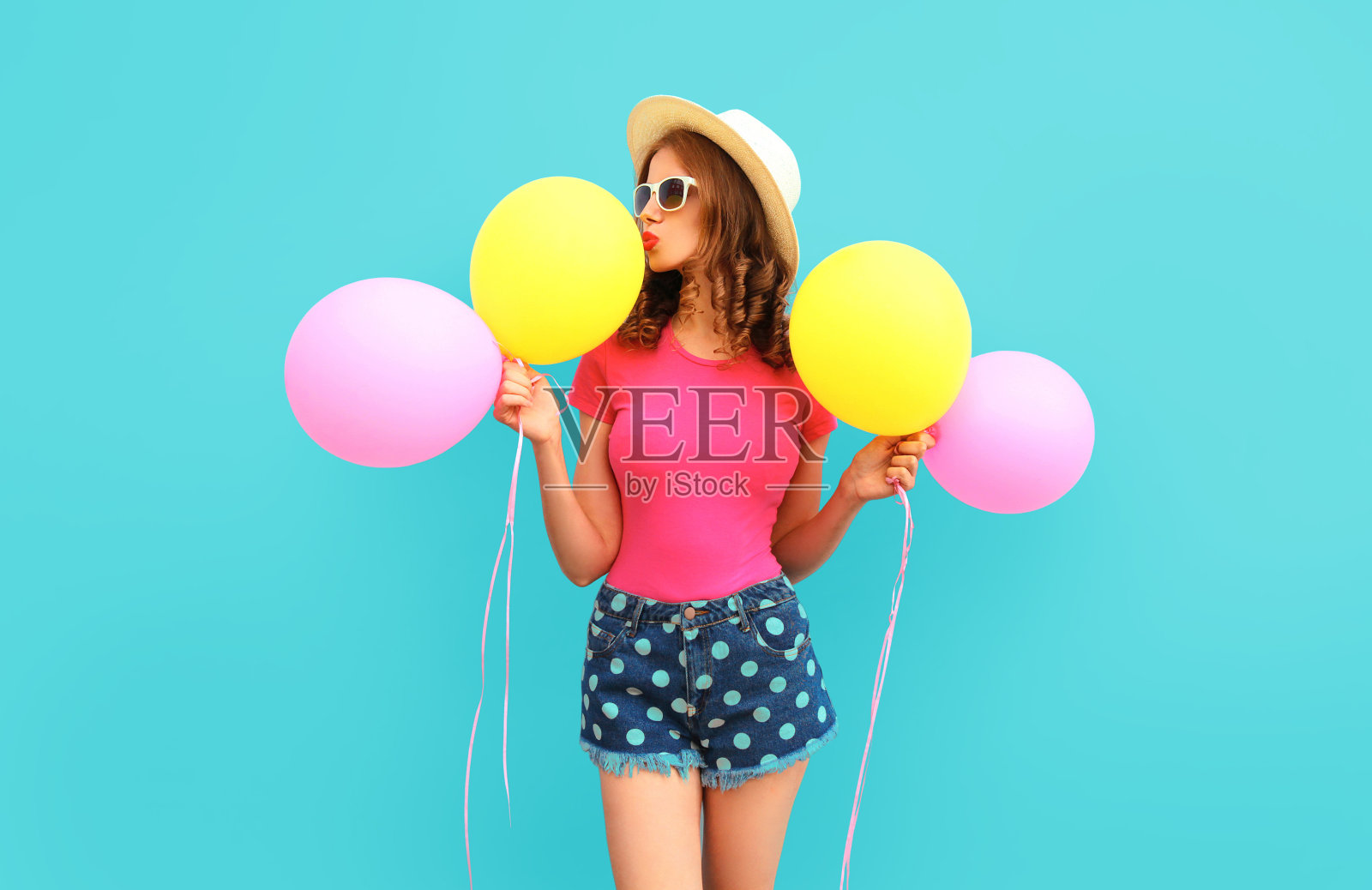 夏天的彩色图像可爱的年轻女子亲吻黄色的粉红色气球穿着短裤和草帽在蓝色的墙壁背景照片摄影图片